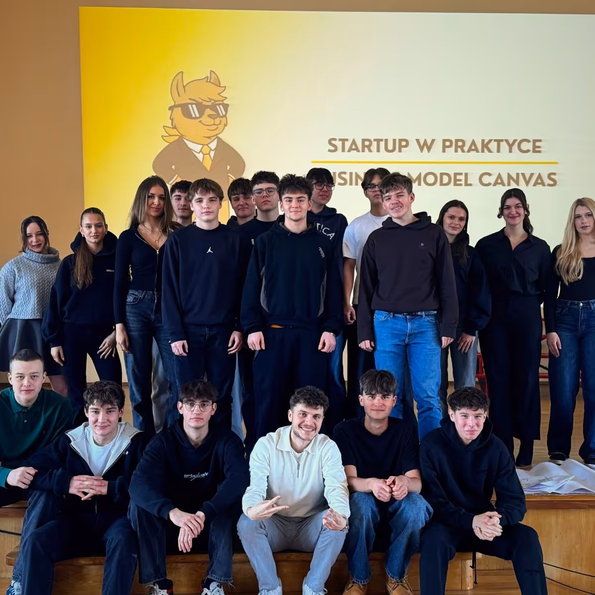 Group of young people posing in front of a yellow screen with a cartoon and the text Startup w praktyce.