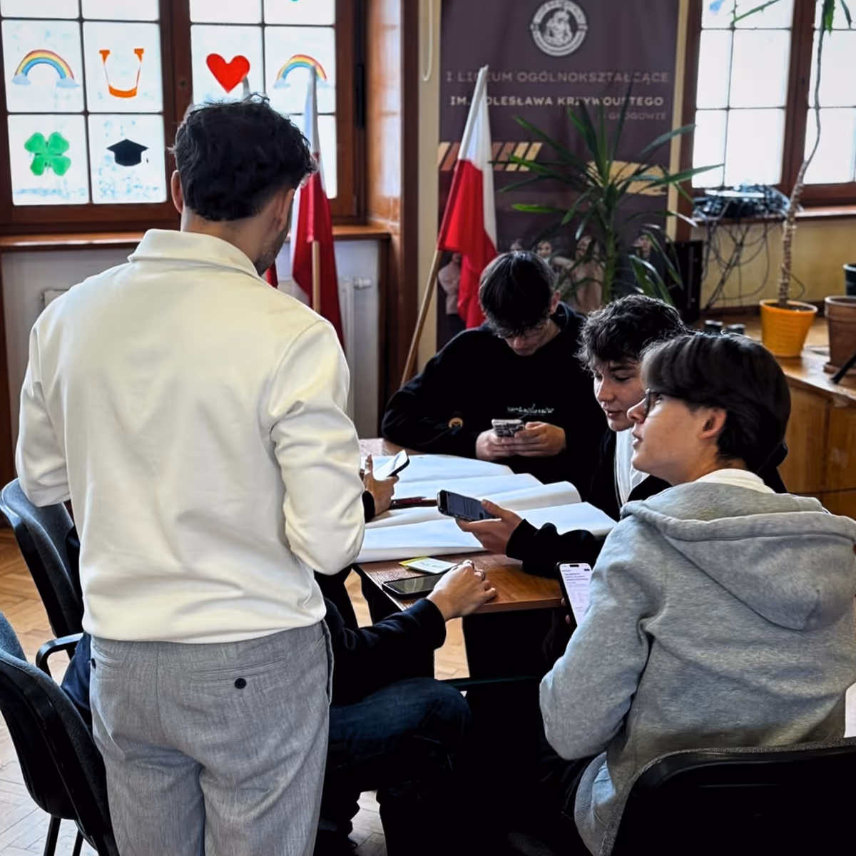 Five young men sitting and standing around a table using smartphones in a room with Polish flags.