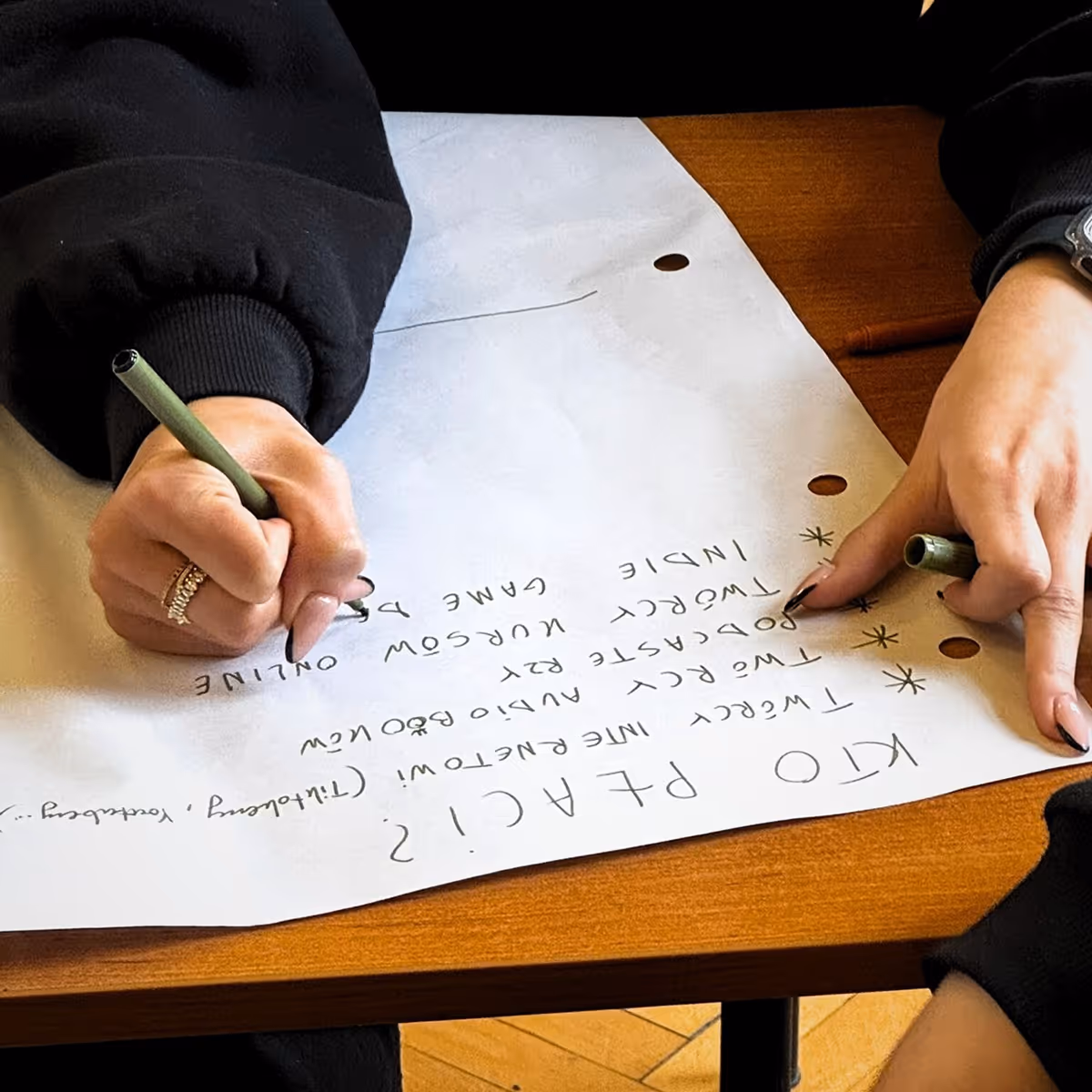 Two hands writing a list of video game titles on white paper on a wooden table.