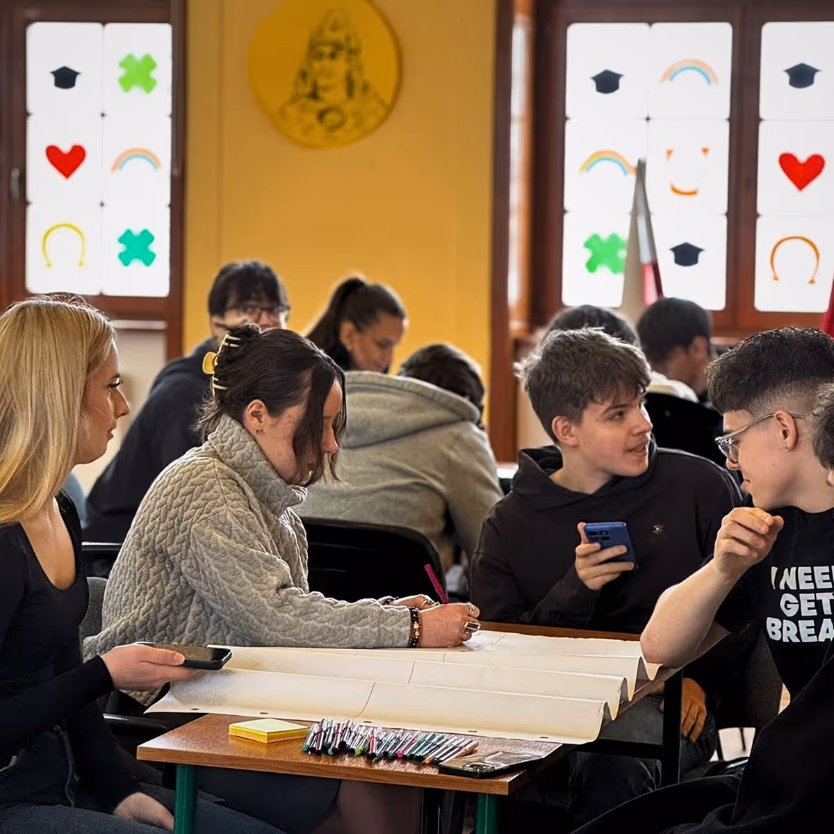 Group of students sitting around a table with pens and large paper, engaged in discussion and writing.