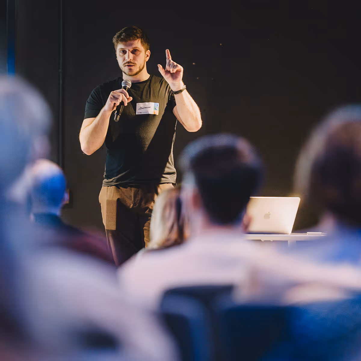 Man speaking into a microphone, gesturing with one hand, facing an audience with a laptop beside him.