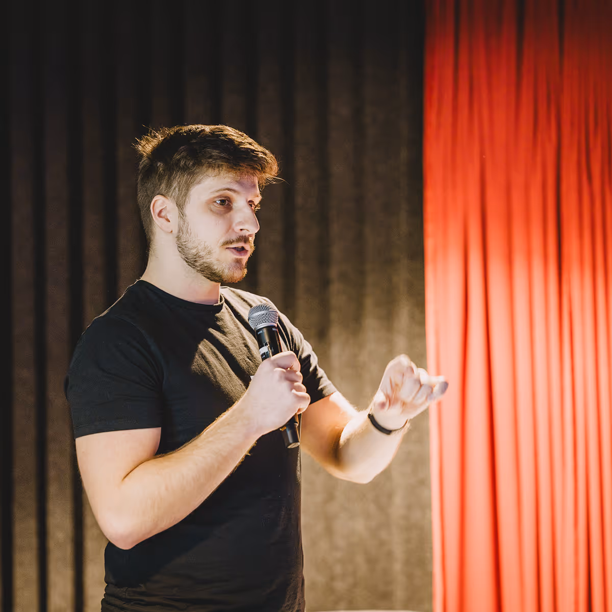 Man in black shirt speaking into a microphone in front of black and red curtains.