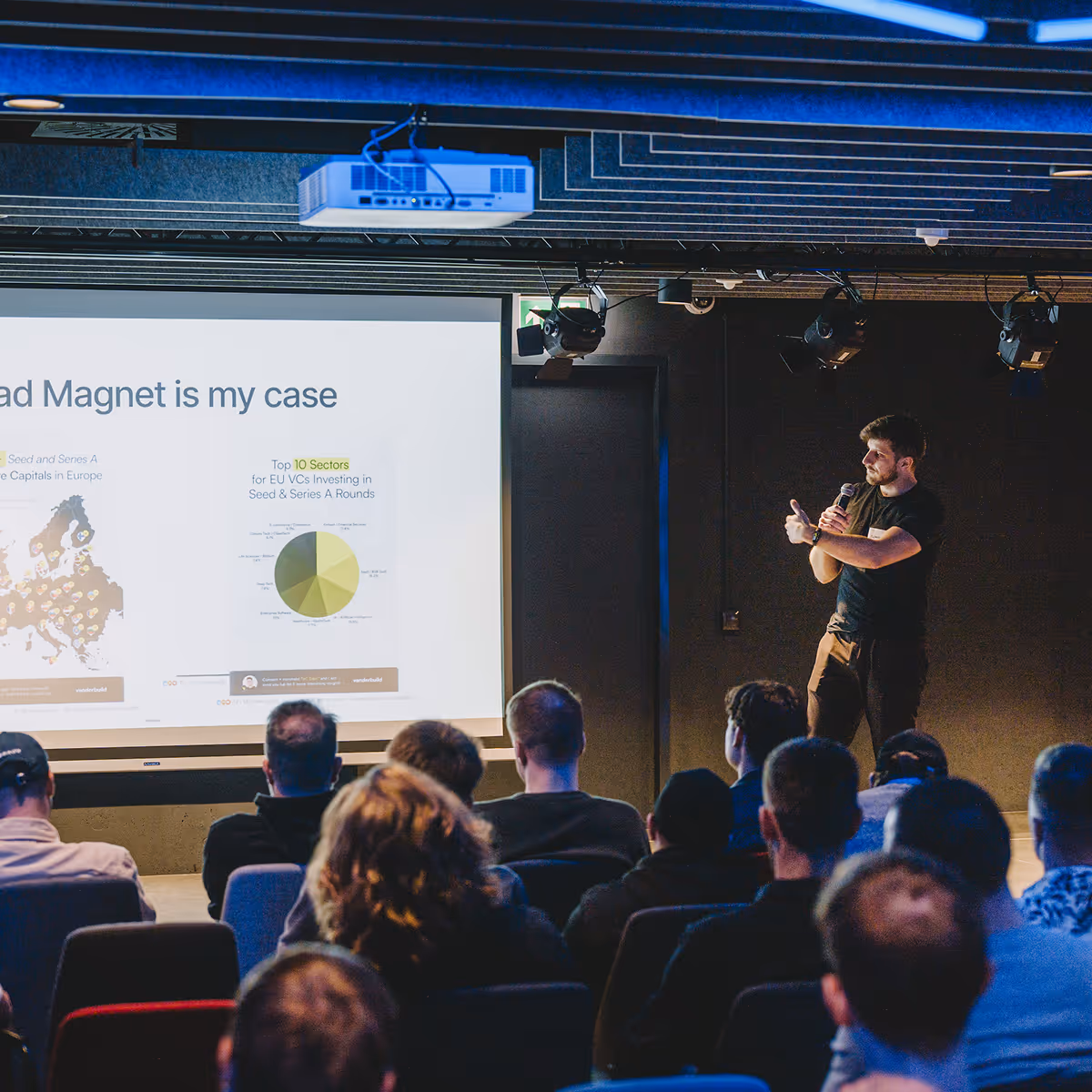 A man gives a presentation with a microphone to an audience facing a screen with charts and a map.