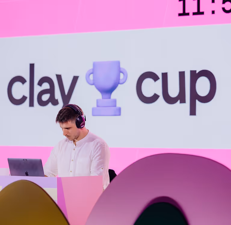Man in white shirt with headphones working on a laptop under a pink Clay Cup sign with a trophy icon.