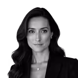 Portrait of a woman with long dark hair wearing a black blazer and a delicate necklace, looking at the camera with a neutral expression.