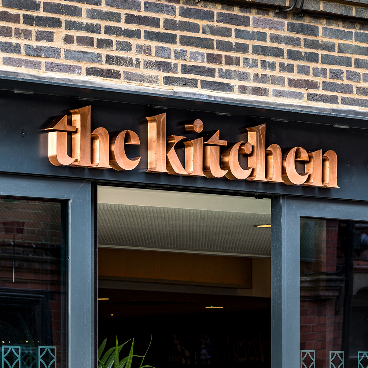 Close-up of the exterior of a restaurant named “The Kitchen,” featuring large copper-colored lettering above a dark-framed entrance set into a brick building.