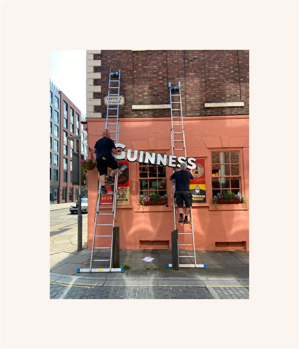 Two workers on ladders install a large Guinness sign on the front of a brick pub.
