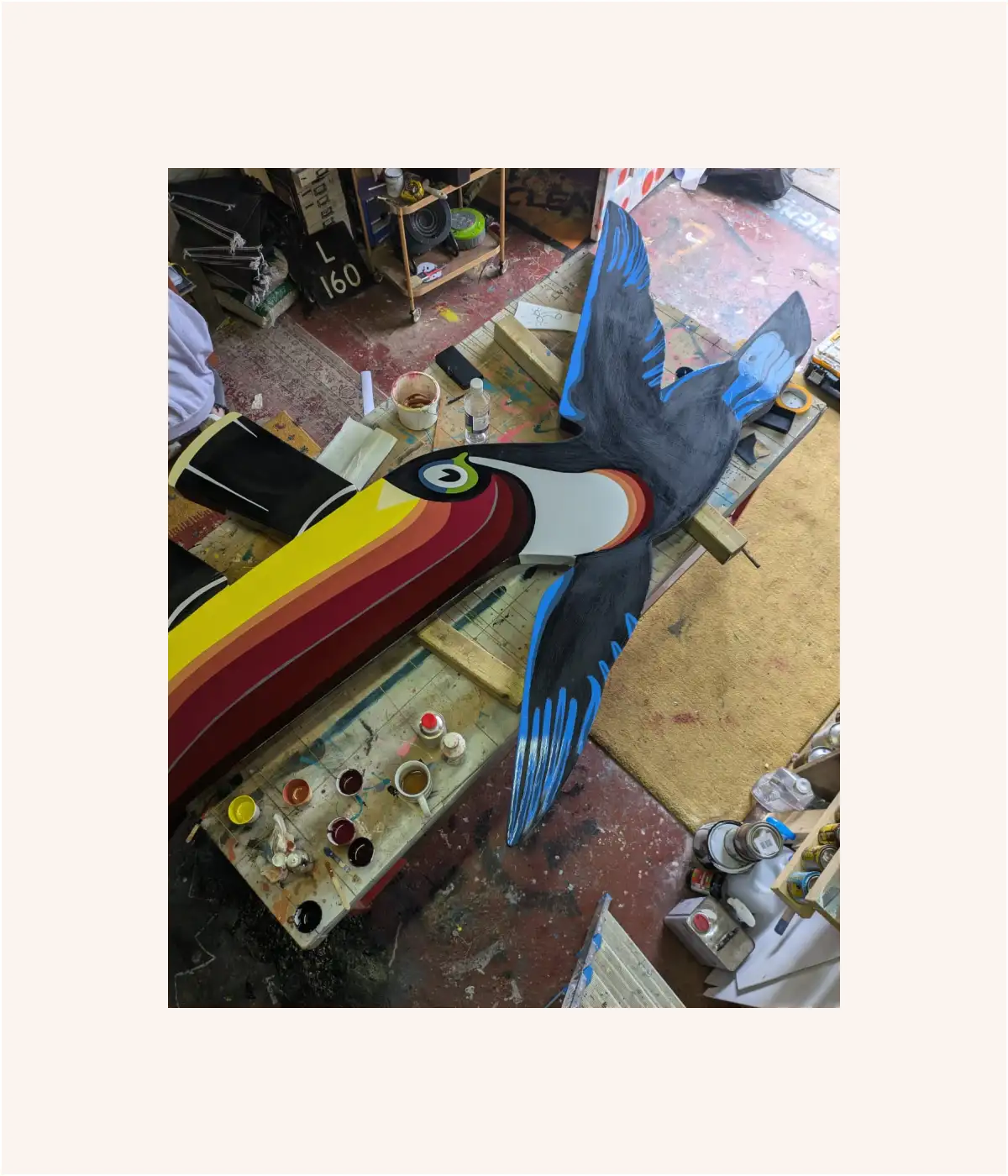 A colorful bird artwork with blue and black wings is being painted in a busy workshop.