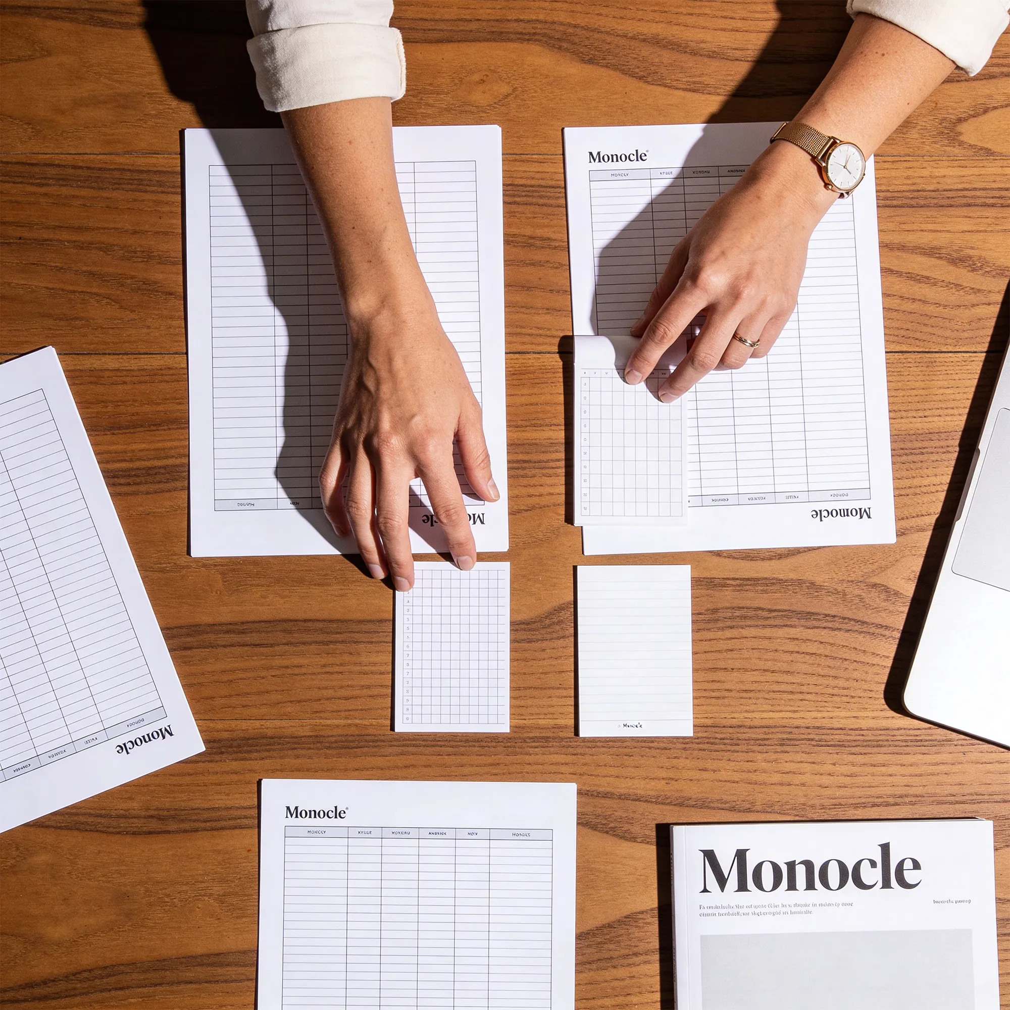 Deux mains rangeant des feuilles d'agenda et des fiches de notes de la marque Monocle sur un bureau en bois, avec un ordinateur portable partiellement visible.