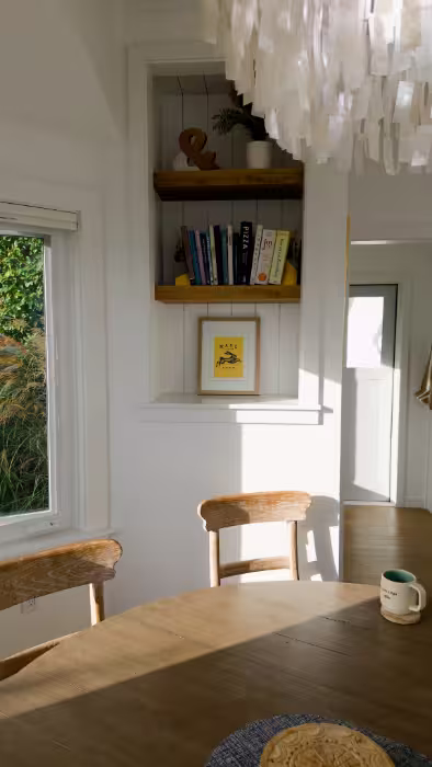 Sunlit kitchen corner with wooden chairs, a mug on the table, framed art, and a bookshelf with books.