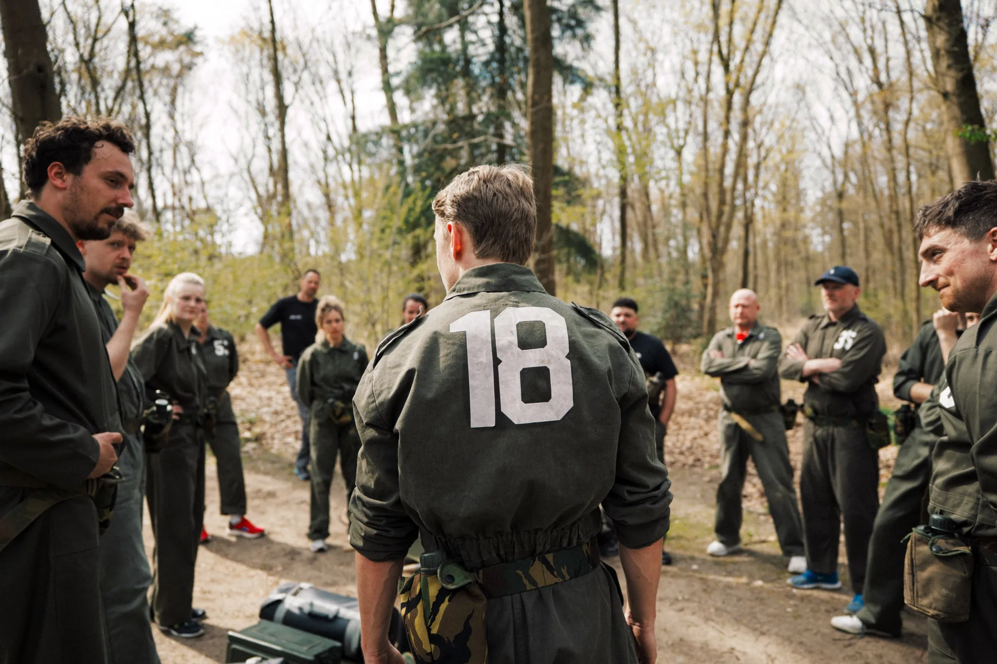 Groep volwassenen in groene trainingen staat buiten in een bos, met een persoon met nummer 18 op zijn rug centraal gezien.