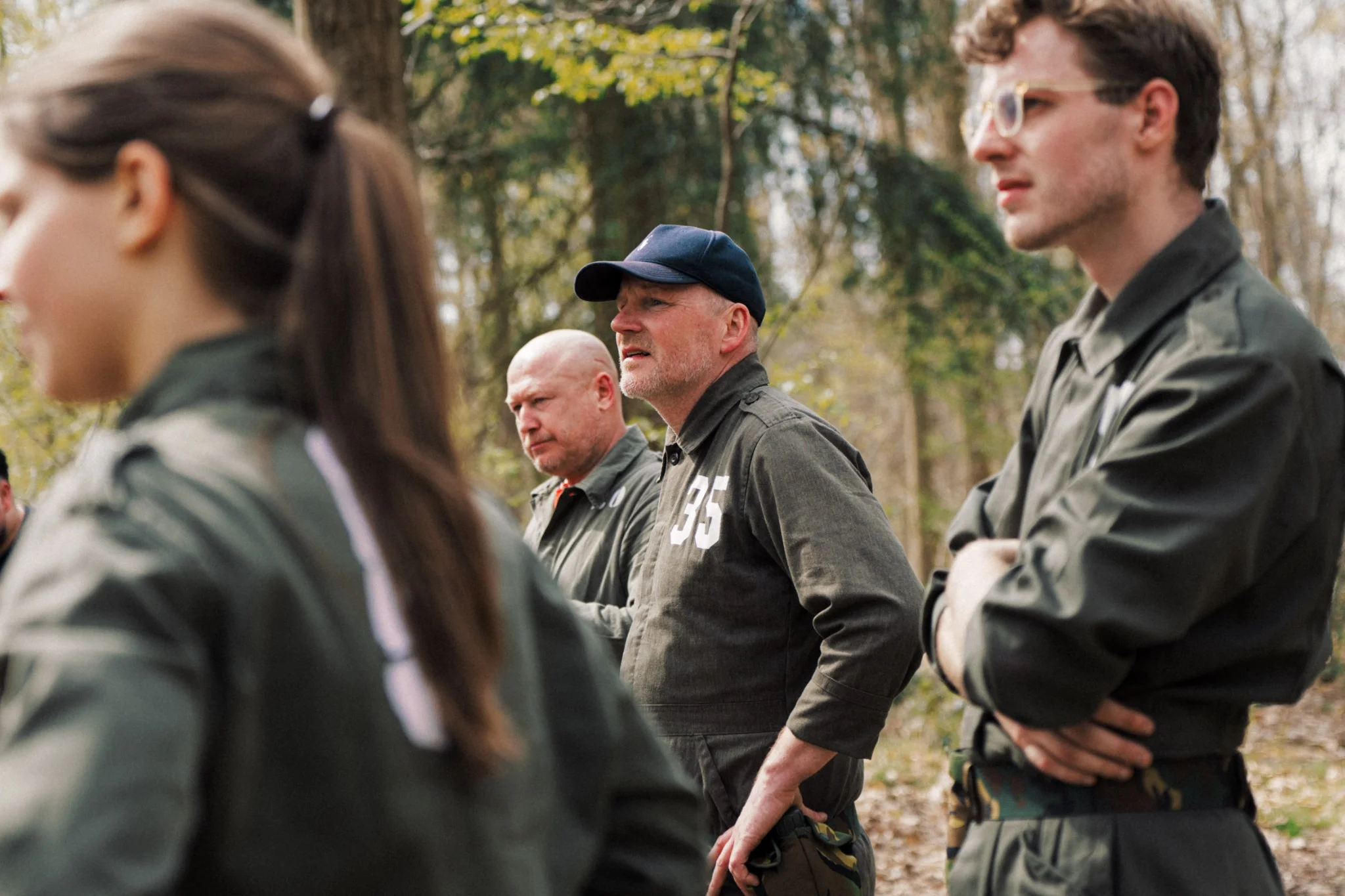 Vier personen in groene militaire kleding staan in een bosrijke omgeving, kijkend naar iets buiten beeld.