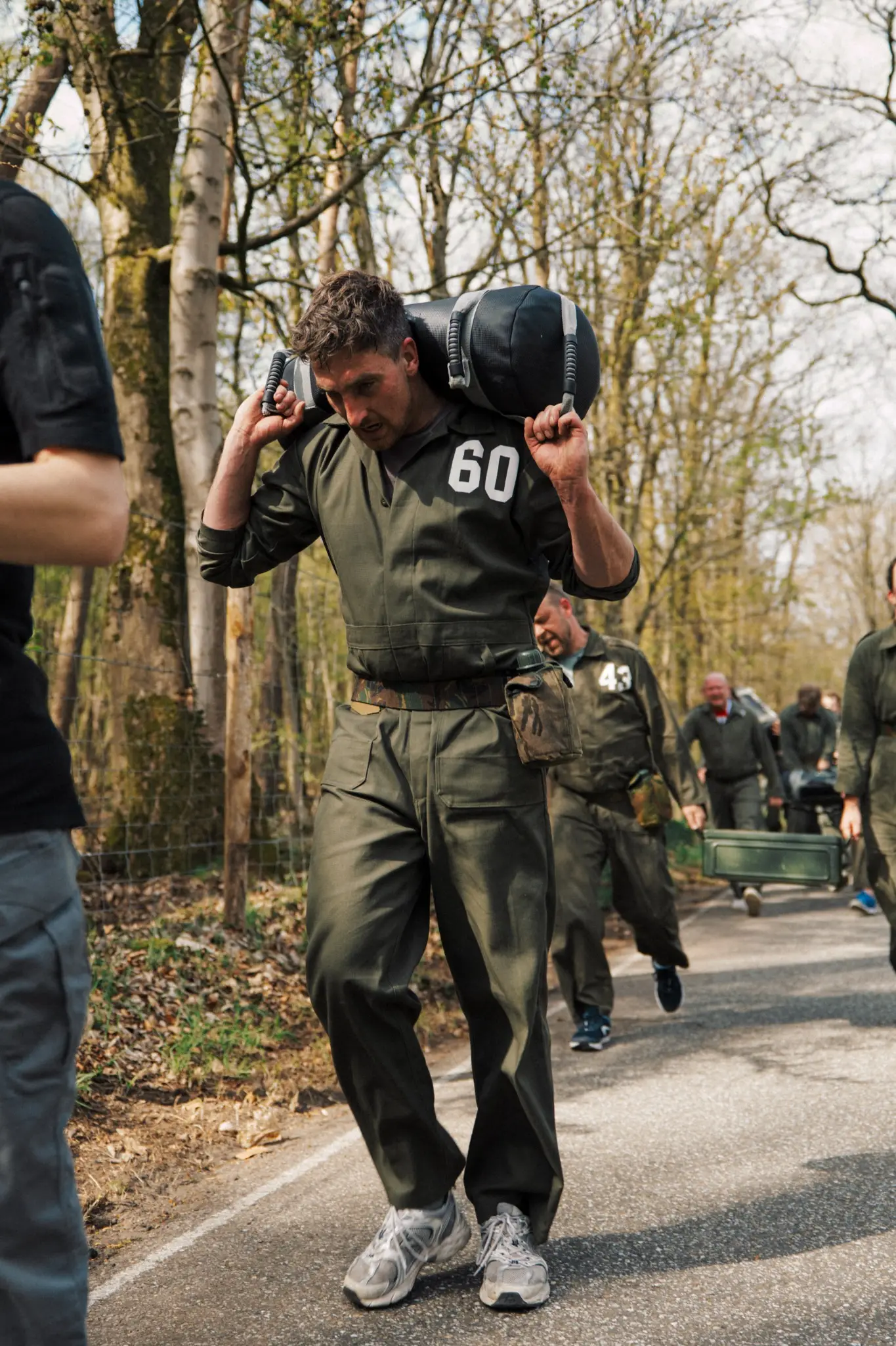 Man in groene overall met nummer 60 draagt een zware zak op zijn schouders tijdens een outdoor training in het bos.