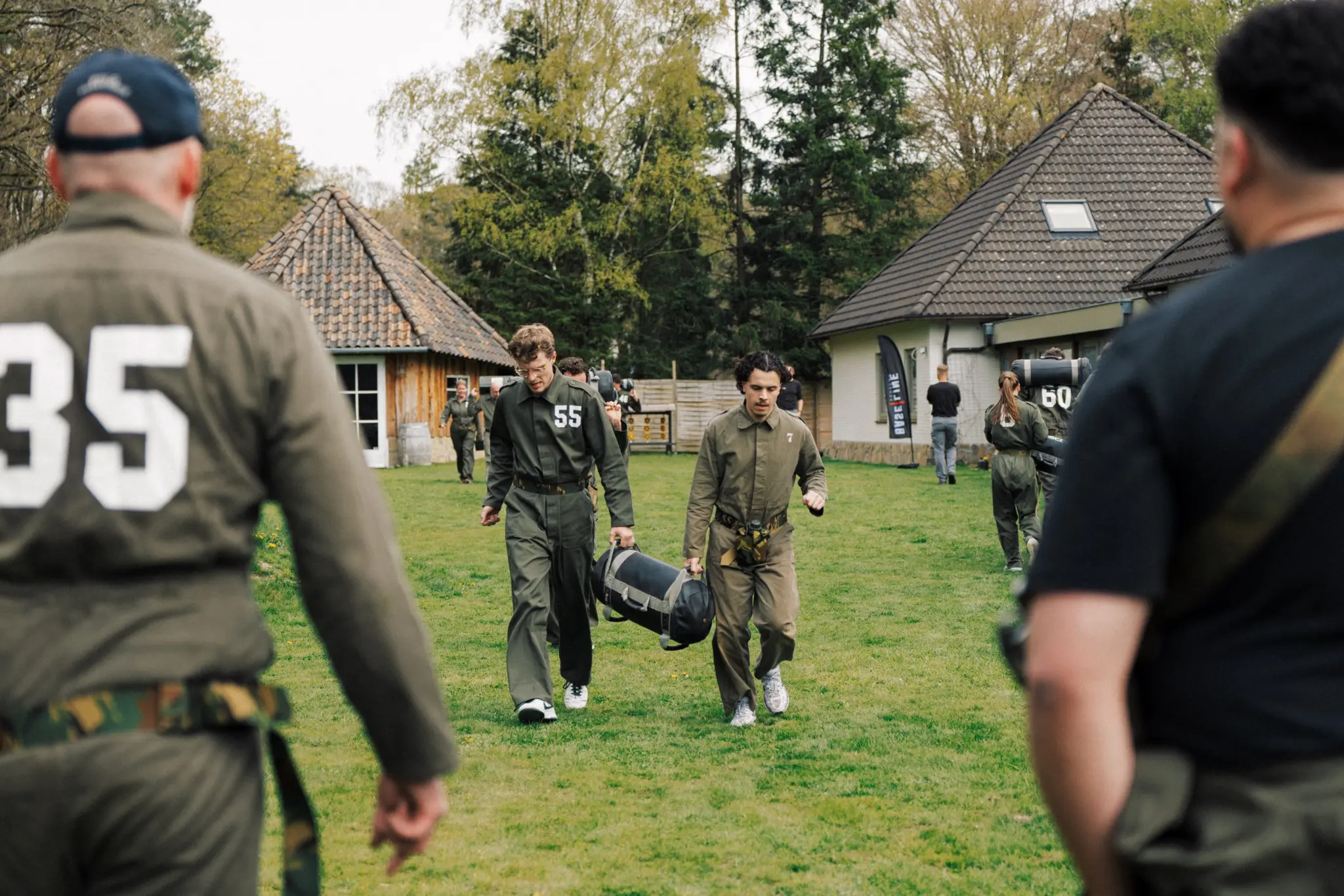 Mensen in militaire kleding lopen op grasveld met houten en stenen huisjes op de achtergrond.