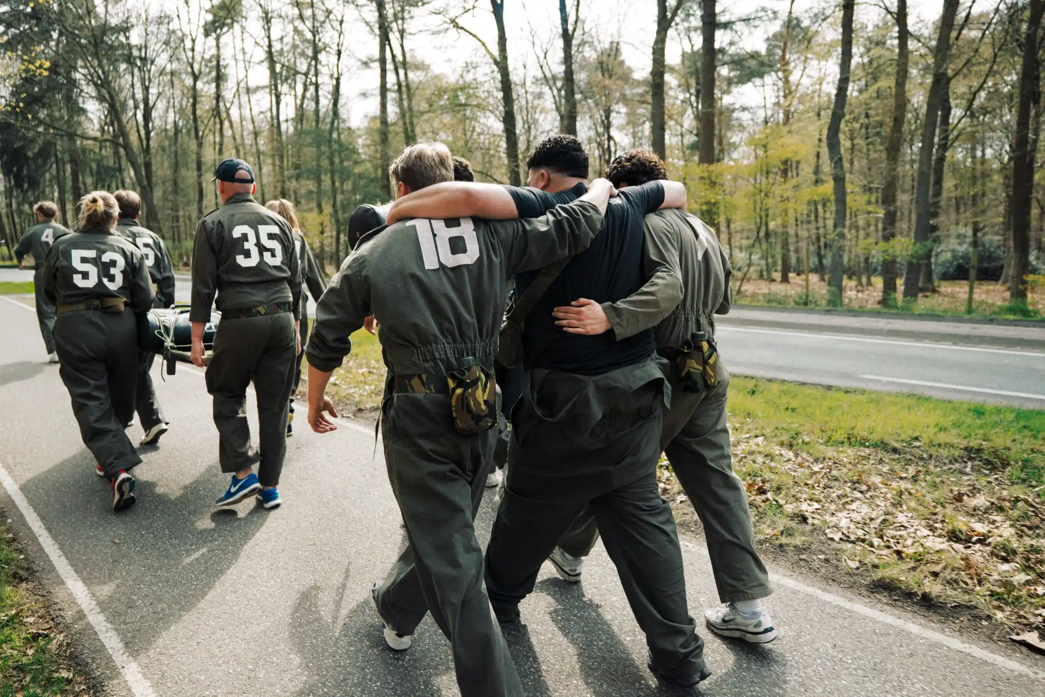 Een groep mensen in militaire kleding wandelen op een weg in een bos, waarbij twee mannen elkaar ondersteunen met hun armen om elkaar heen.