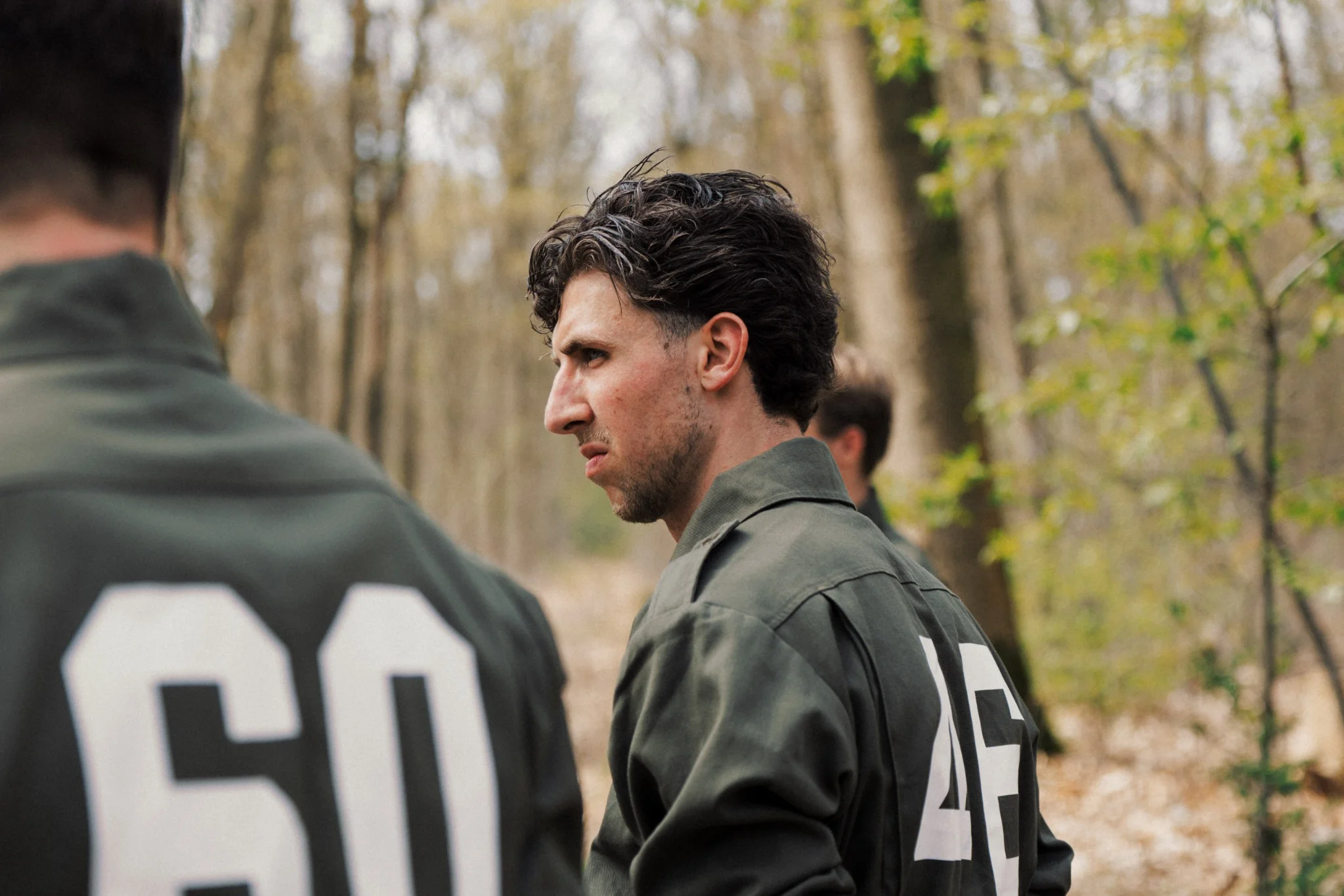 Man met een groene jas met het nummer 45 op de rug staand in een bosrijke omgeving, omringd door anderen met gelijkaardige jassen.