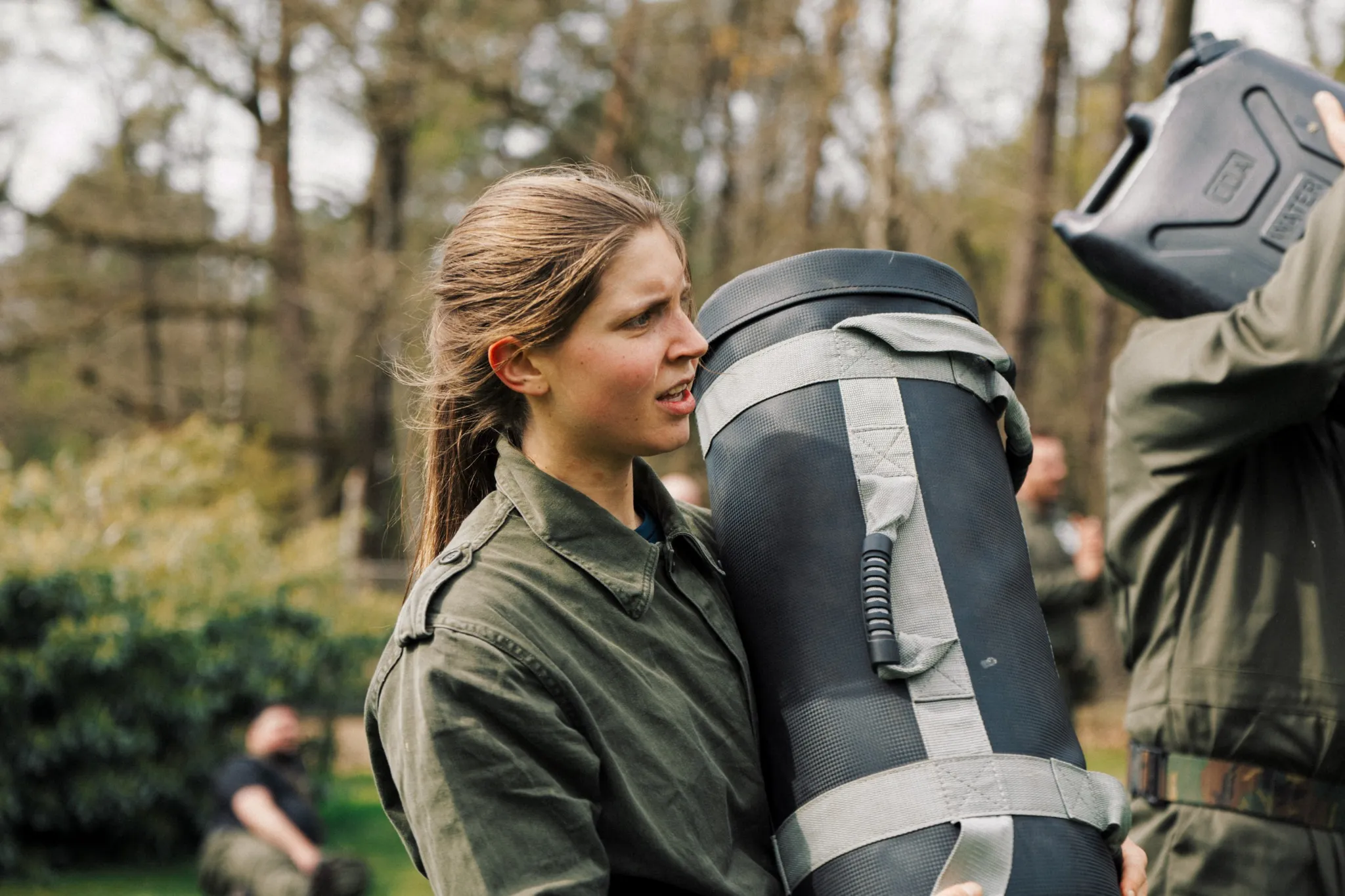 Vrouw in groen militair uniform draagt een opgerolde mat buiten in een bosrijke omgeving.