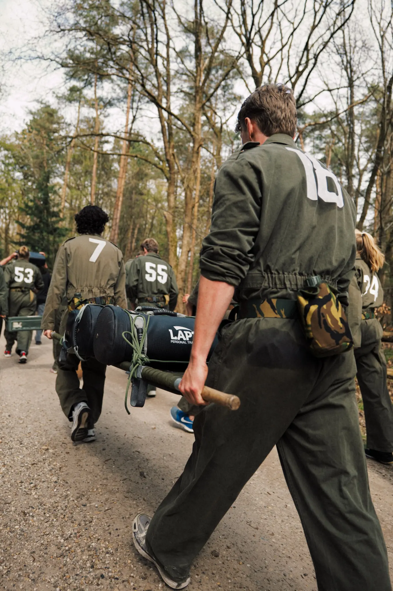 Een groep mensen in groene militaire kleding met nummers op hun rug wandelt over een bosweg, waarbij één persoon een fitness-zak op een stok draagt.