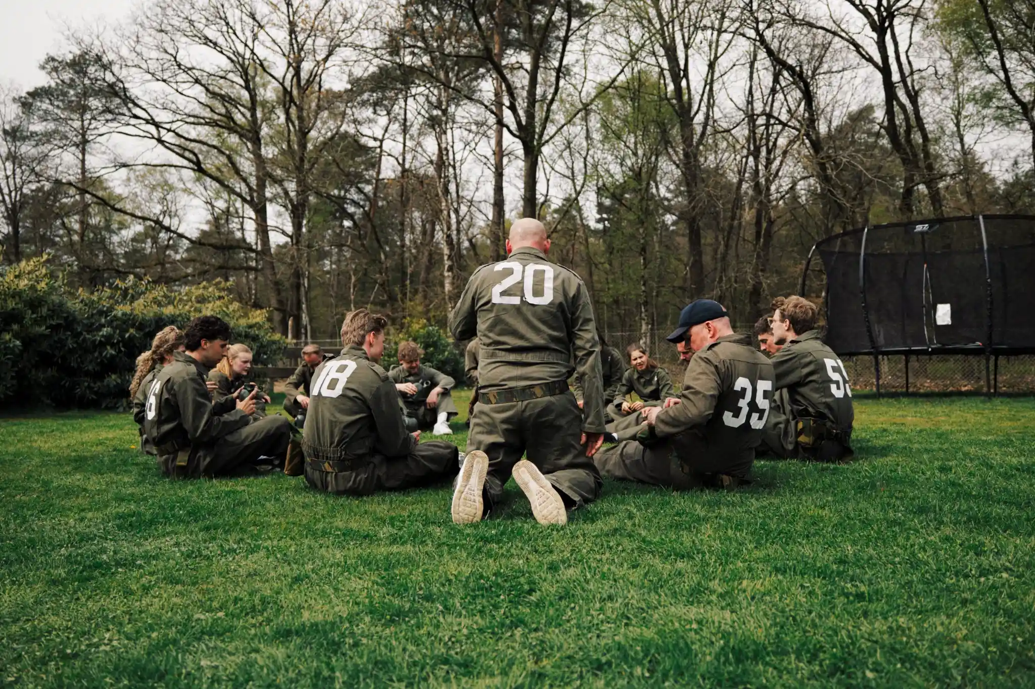Groep jonge mensen in militaire kleding zittend in een cirkel op grasveld met bomen op de achtergrond.