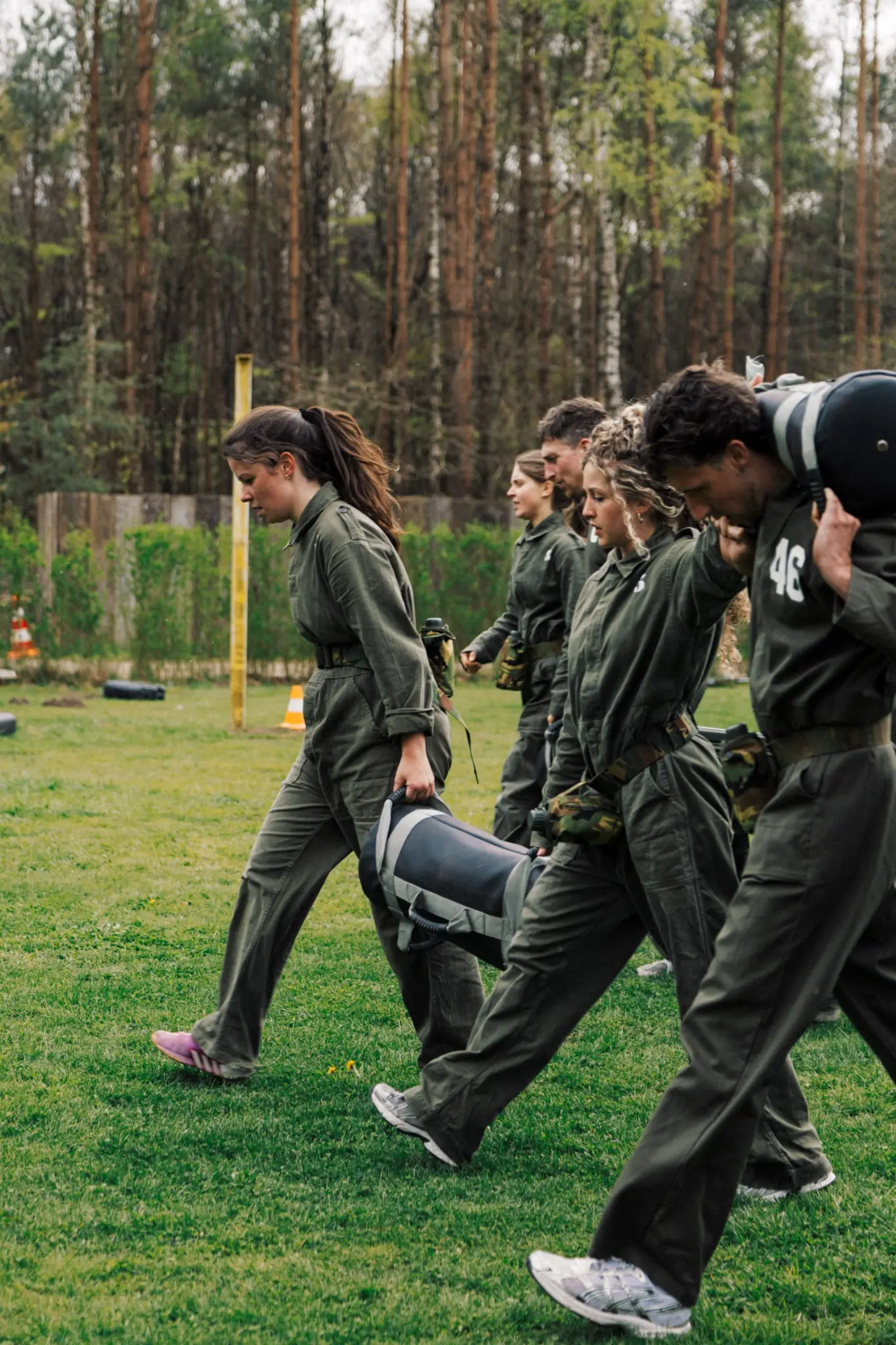 Groep mensen in militaire uniformen die samen sjouwen met zware tassen buiten op een grasveld.