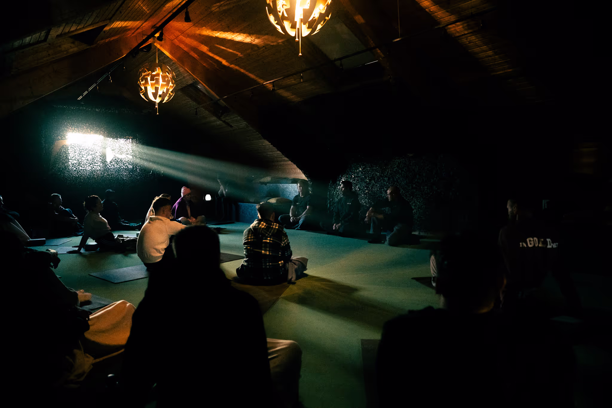 Groep mensen die mediteren in een schemerige ruimte met houten plafond, verlicht door zonlicht dat door een raam schijnt.