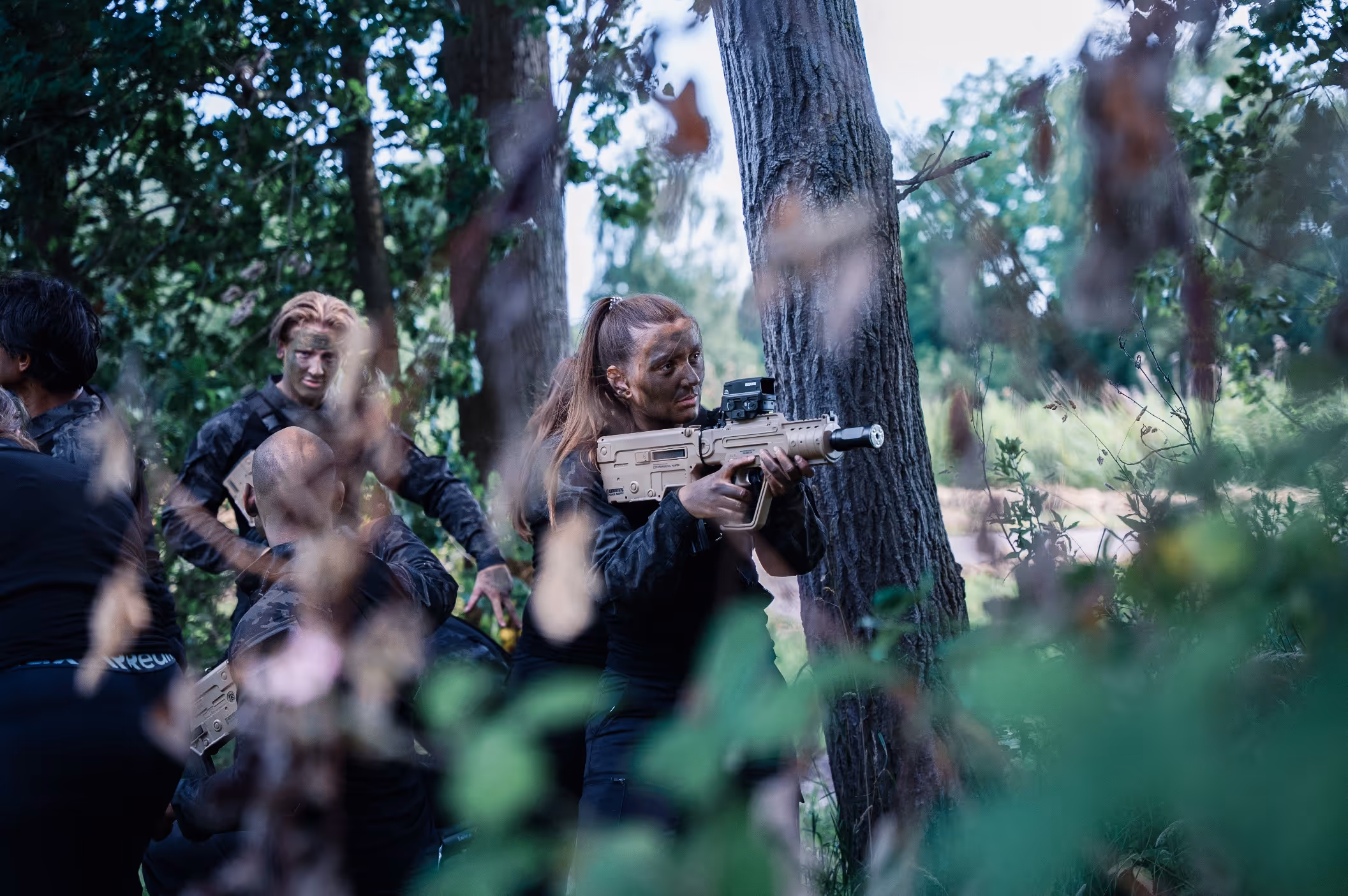 Vrouw met camouflerende gezichtsverf houdt een geweer vast terwijl ze in het bos achter een boom staat.
