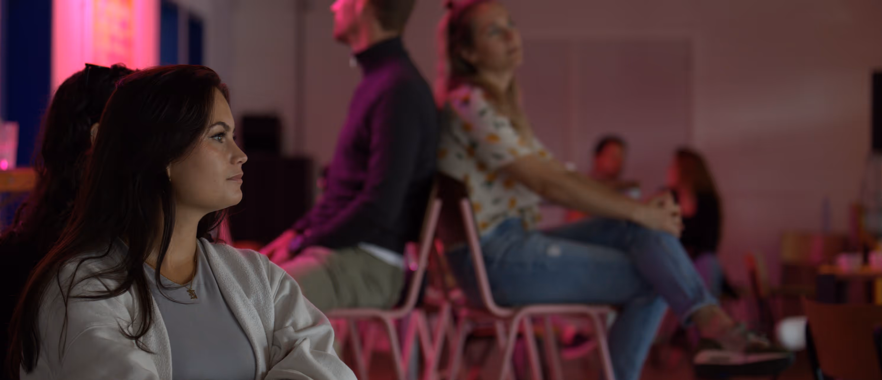 Vrouw met donker haar en lichte trui zittend en luisterend in een zaal met andere mensen op stoelen.