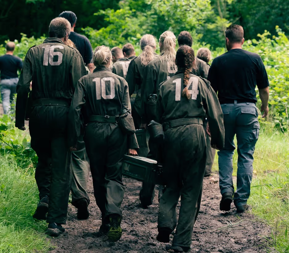 Groep mensen in groene uniformen met nummers op hun rug lopen door een bos op een modderig pad.