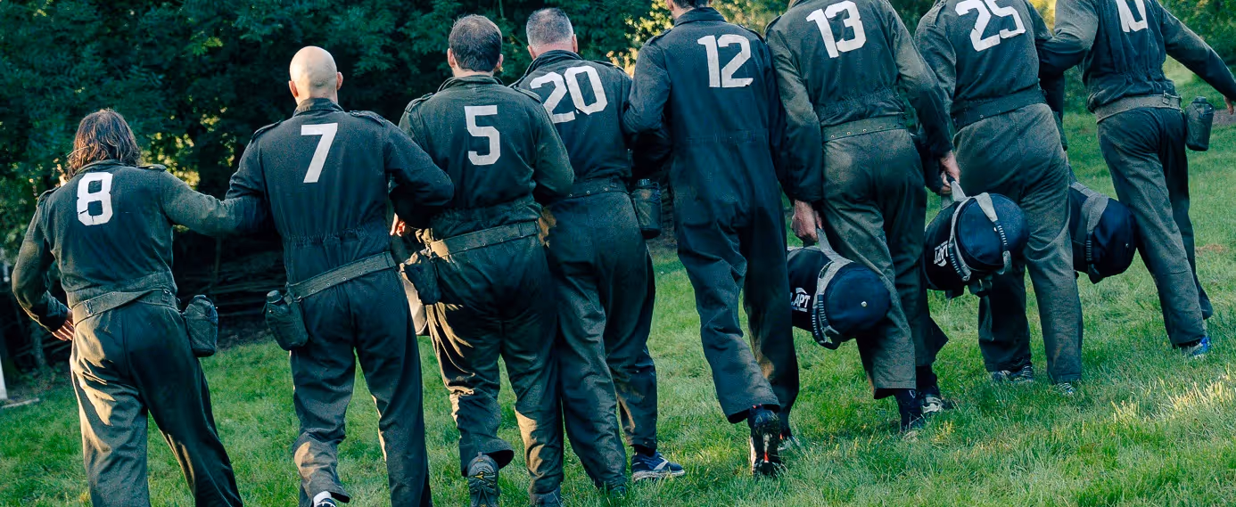 Tien mannen in groene overallpakken met rugnummers van 5 tot 25, lopen samen op gras met helmen in hun handen.