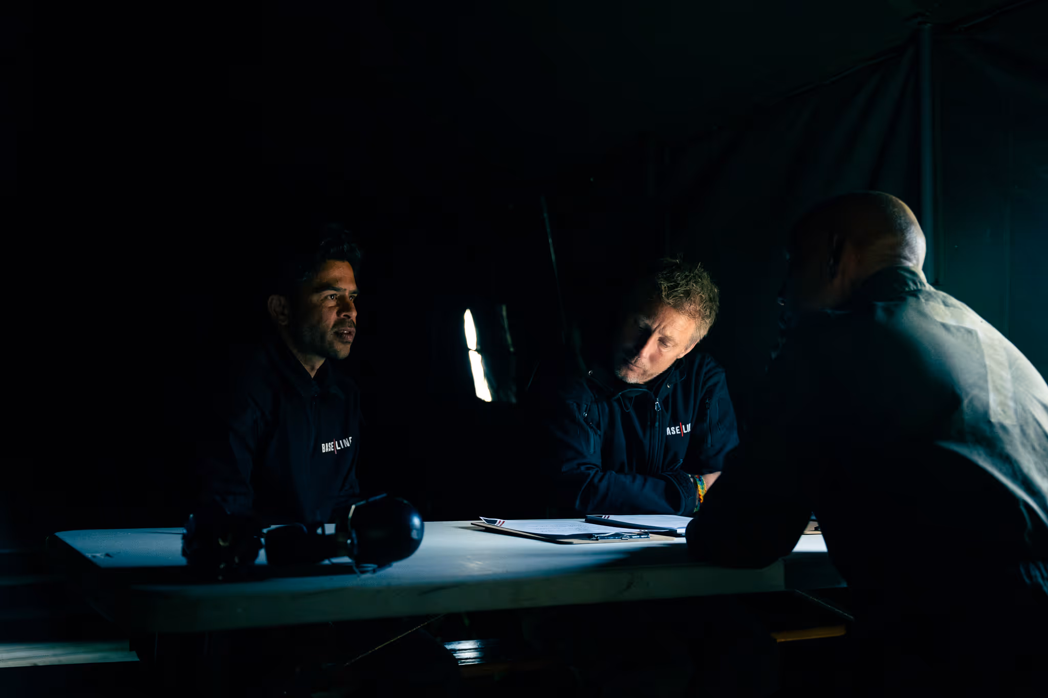 Drie mannen zitten in een donkere ruimte aan een tafel met papieren, alleen subtiel verlicht door een zwak licht.