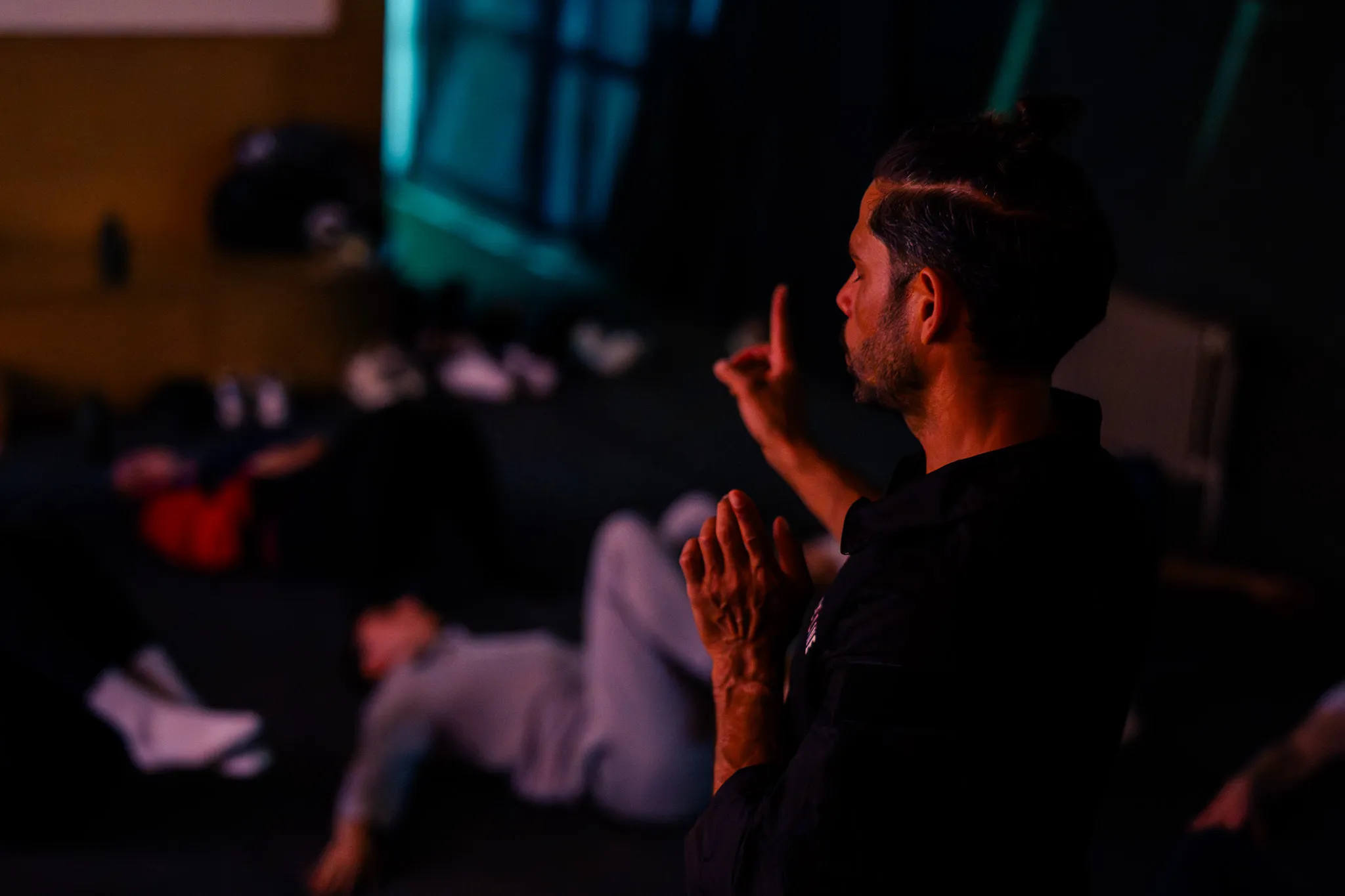 Man met opgetrokken haar die met zijn handen een meditatie- of aanwijzingshouding maakt, met ontspannen mensen op de achtergrond.