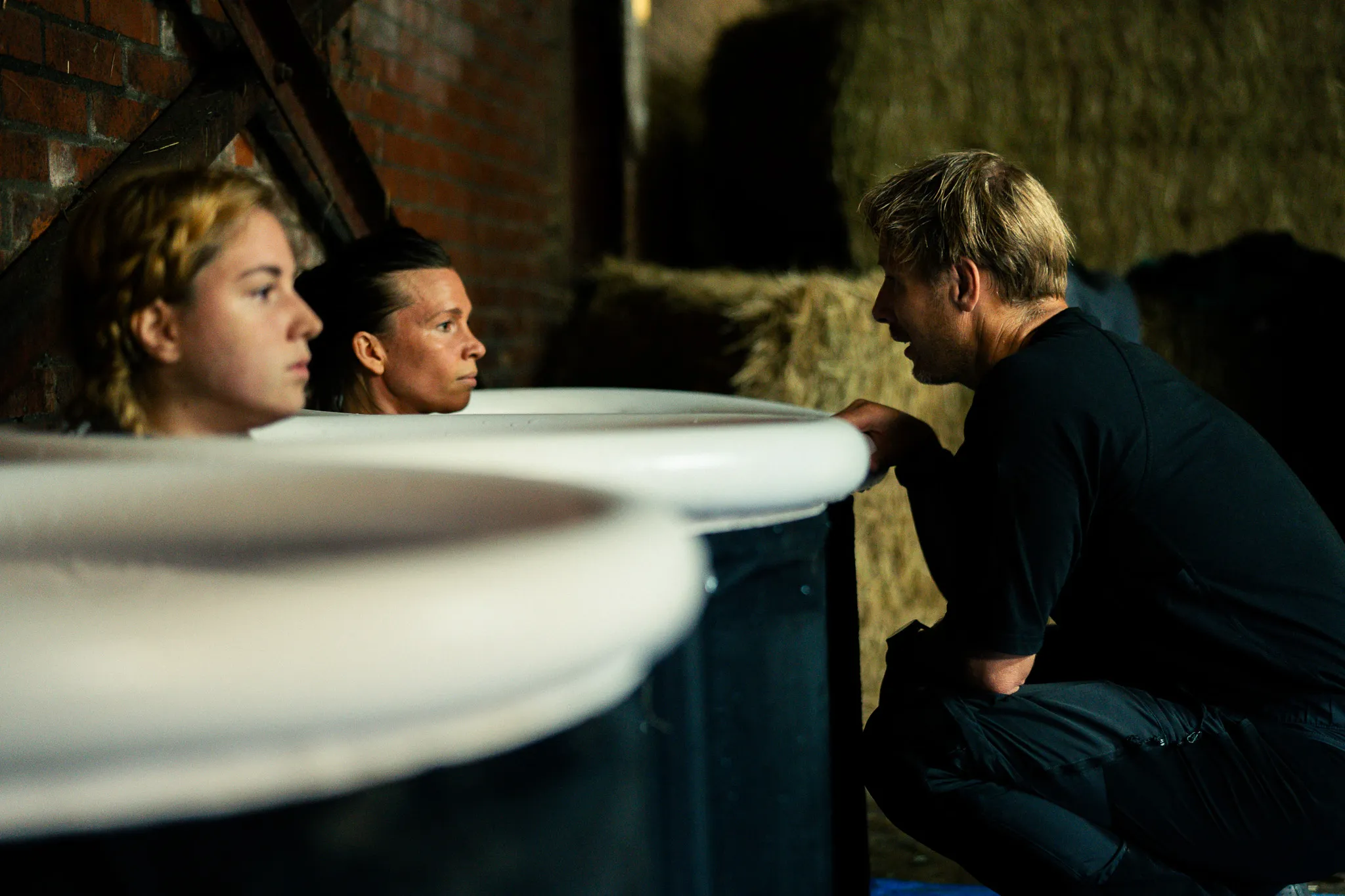 Twee vrouwen zitten in grote baden met een man die voor hen knielt en met hen praat in een donkere schuur met hooibalen op de achtergrond.