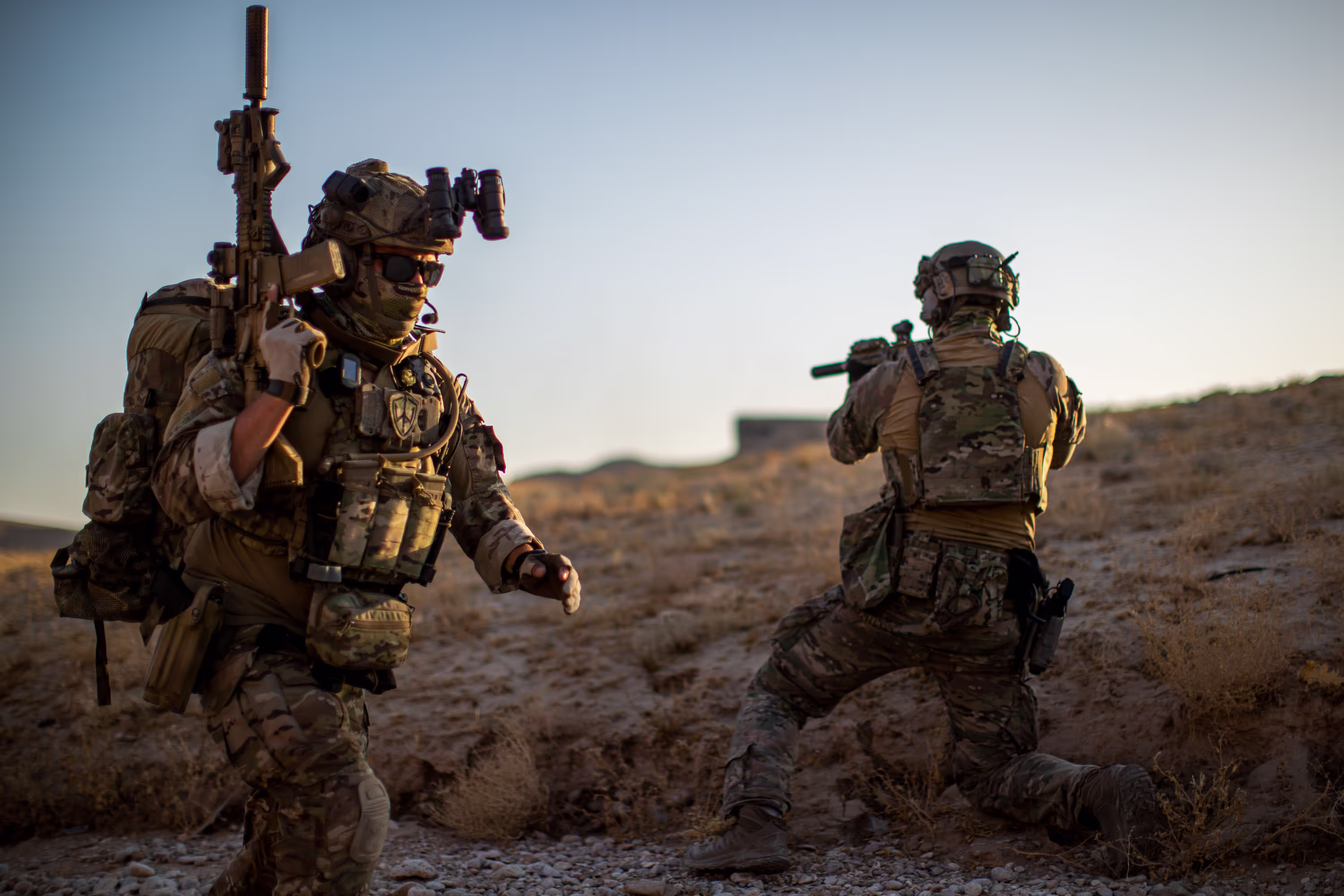 Twee zwaar bewapende soldaten in camouflage in een droog, rotsachtig landschap bij zonsondergang.