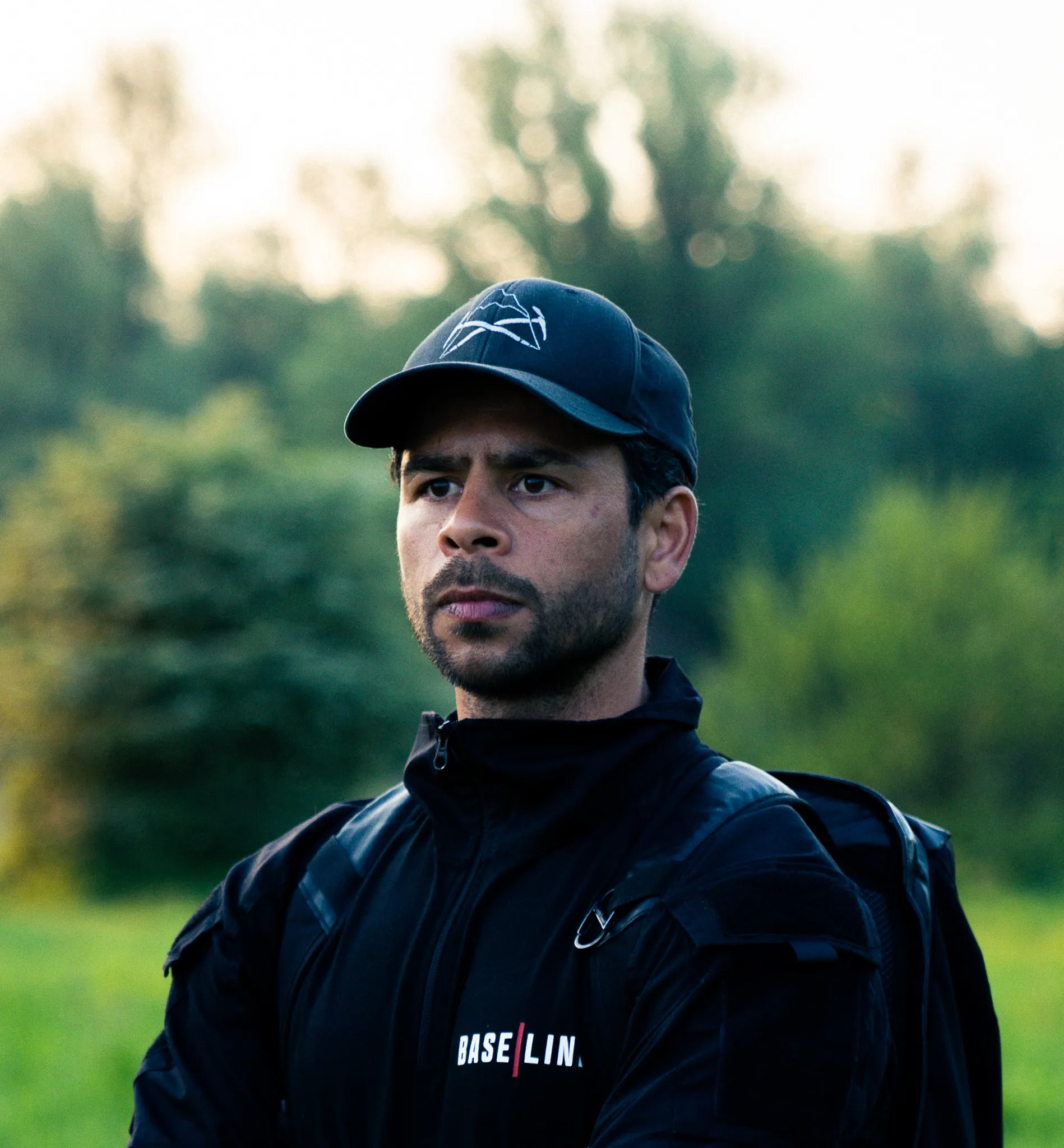 Man met een zwarte pet en jas met de tekst 'BASE|LIN' staand in een groene buitenomgeving.