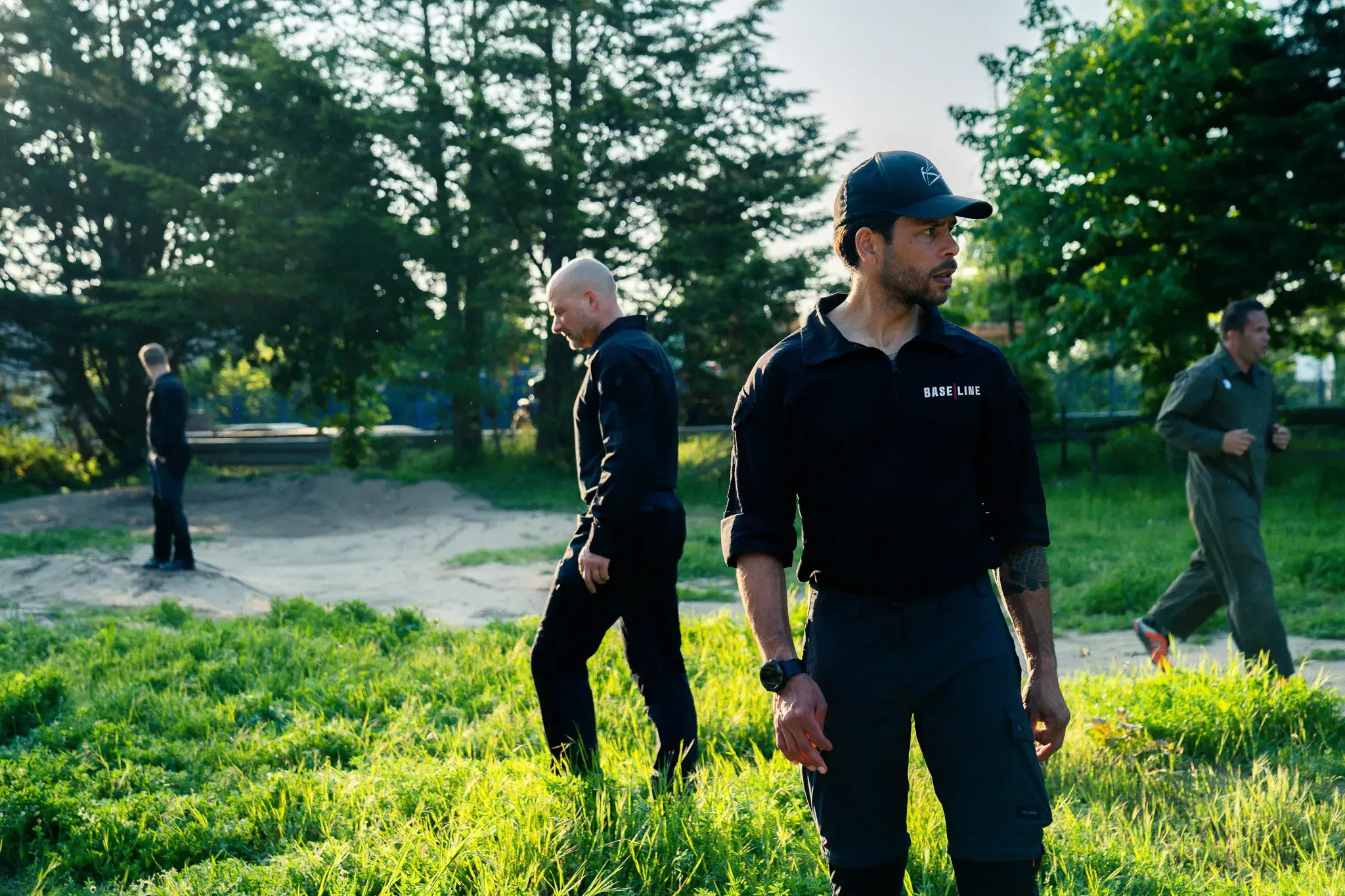 Vier mannen in outdoor kleding in een grasveld, een draagt een zwarte pet met opschrift 'BASE LINE'.