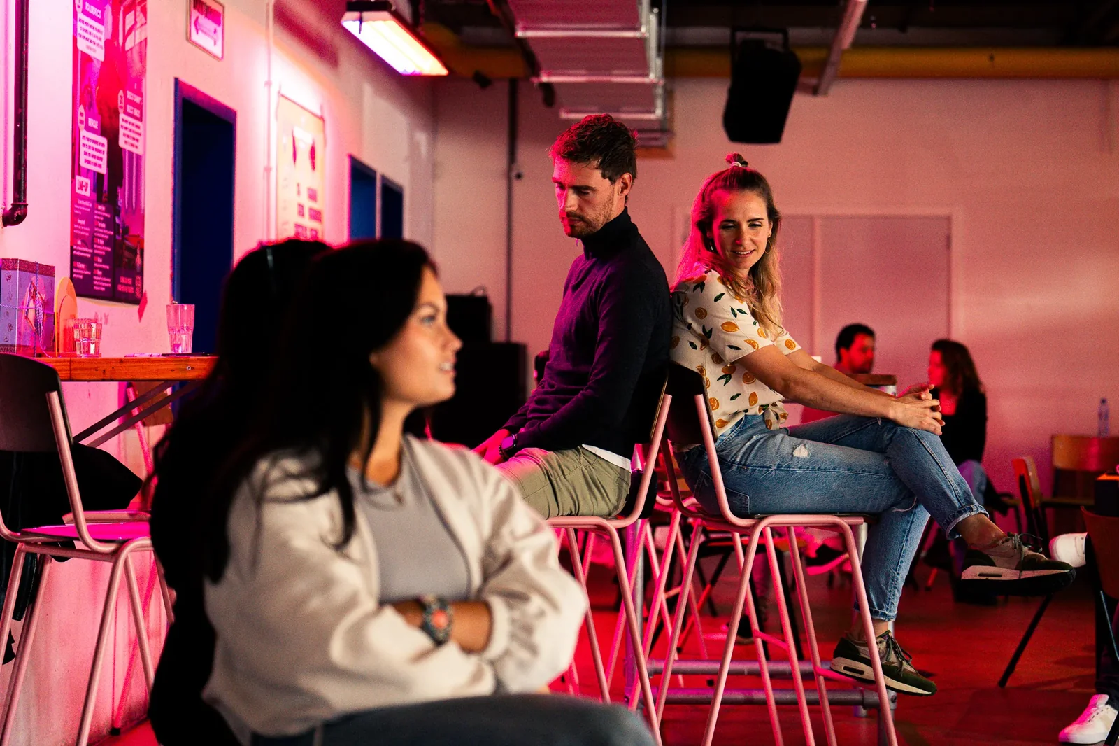 Mensen zitten op hoge krukken in een sfeervolle ruimte met roze verlichting, sommigen praten en lachen.