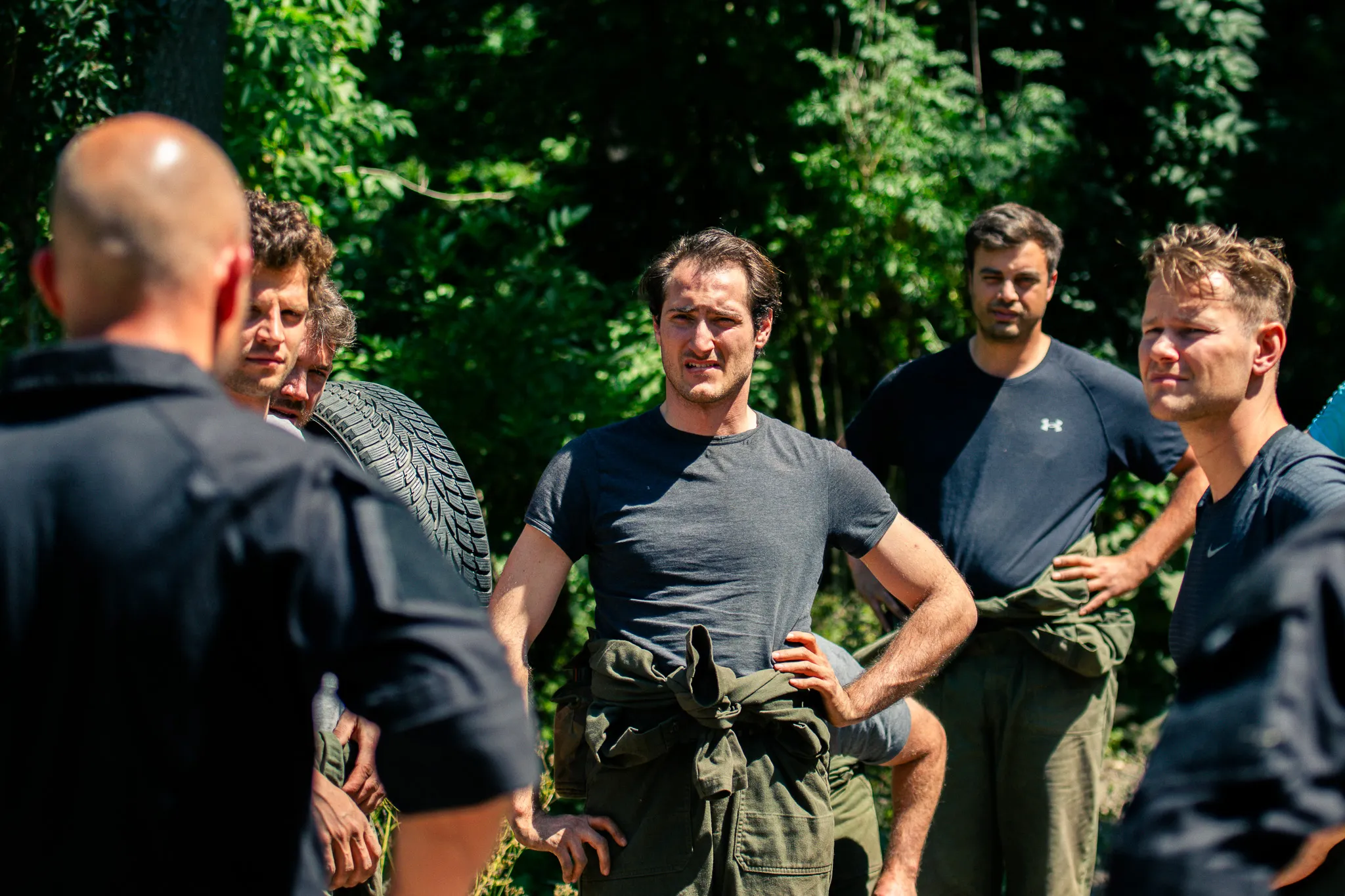 Een groep mannen staat buiten in een bosrijke omgeving met de handen in de zij, sommigen dragen trainingskleding en werkkleding.