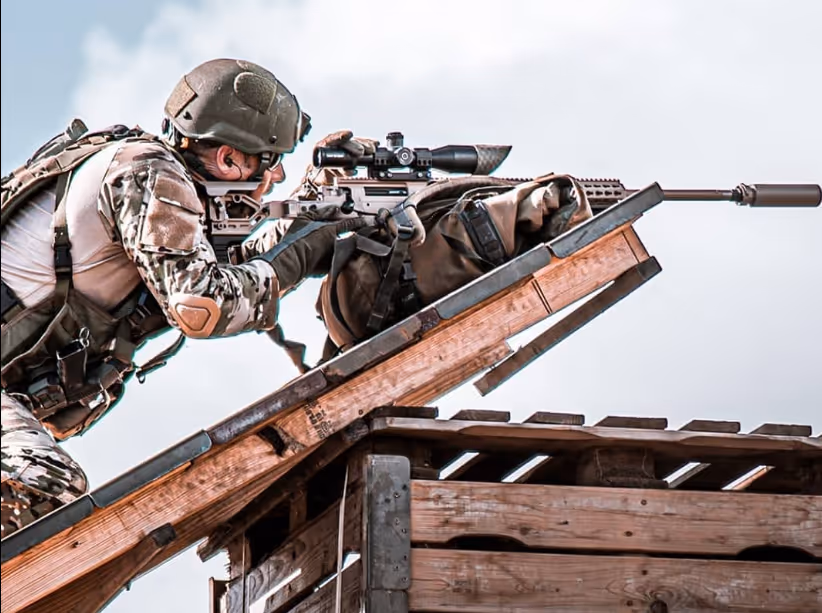 Soldaat in camouflage met helm richt op een sluipschuttersgeweer op een houten platform.