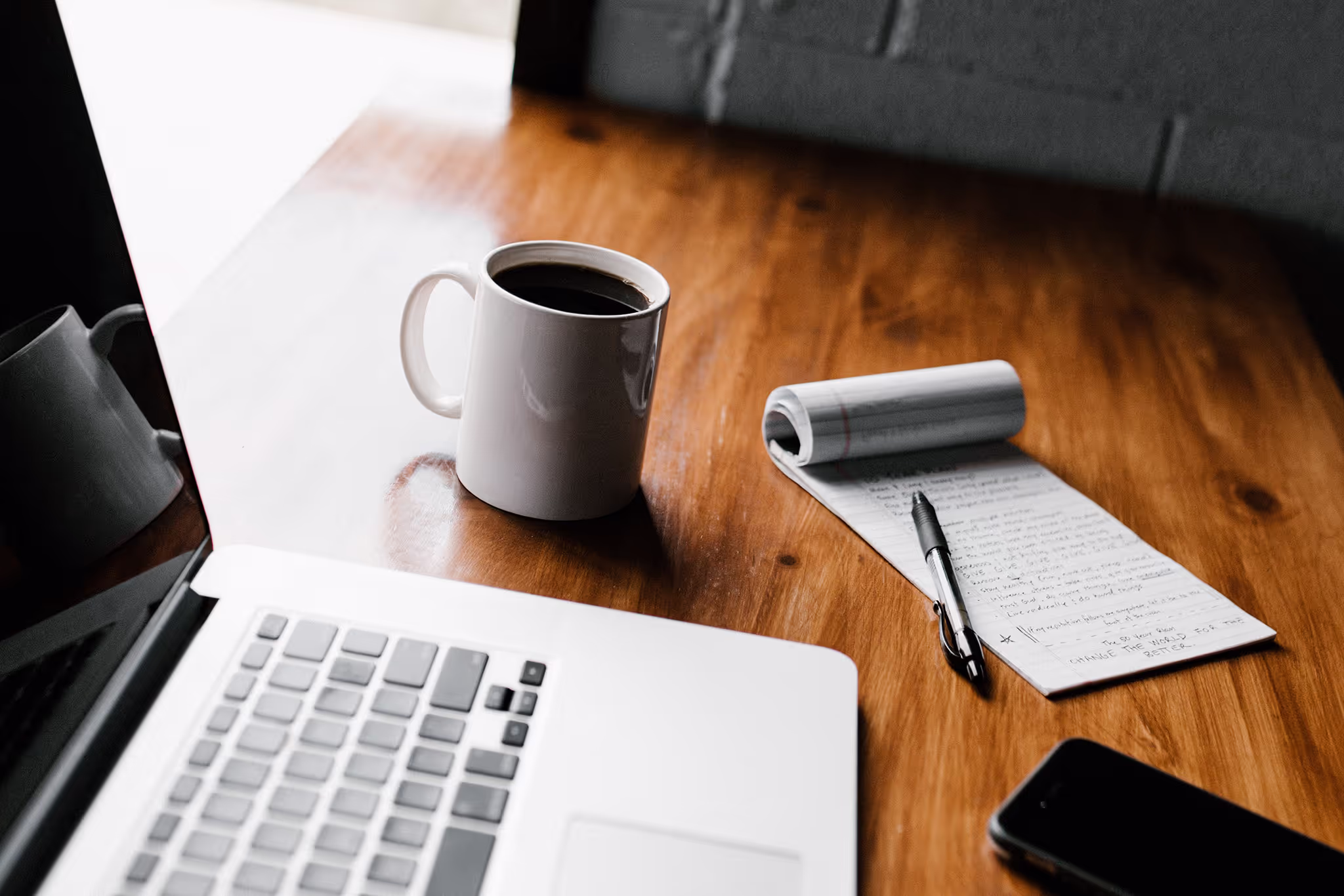 Bureautafel met een open laptop, een witte koffiemok, een notitieblok met pen en een smartphone.