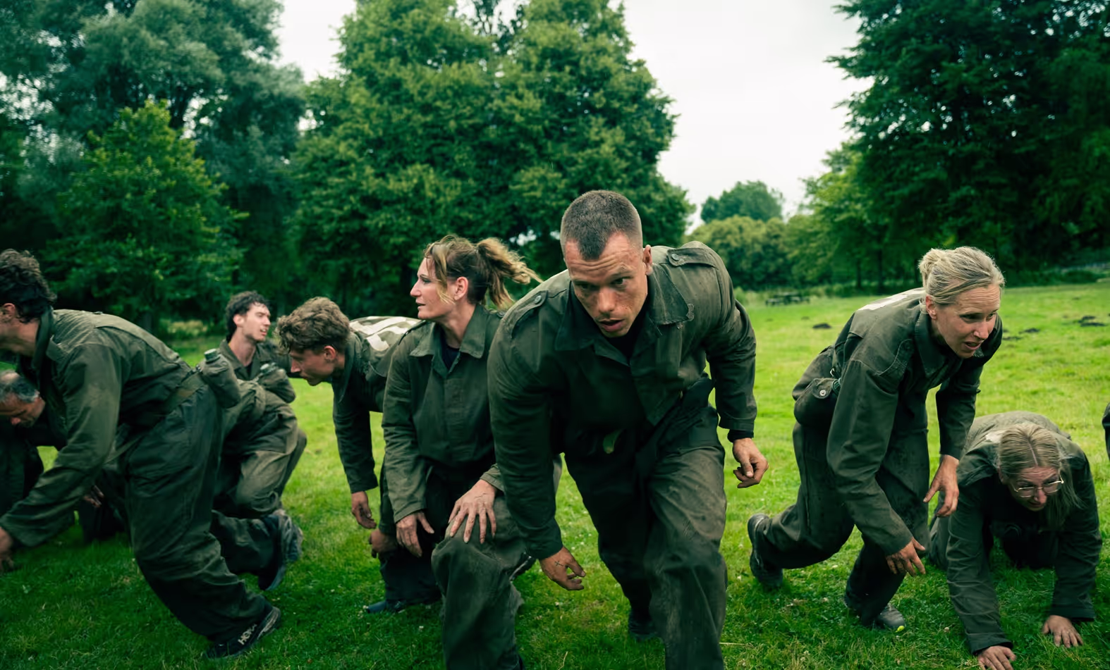 Groep mensen in militaire kleding kruipt op handen en knieën over grasveld in een bosrijke omgeving.