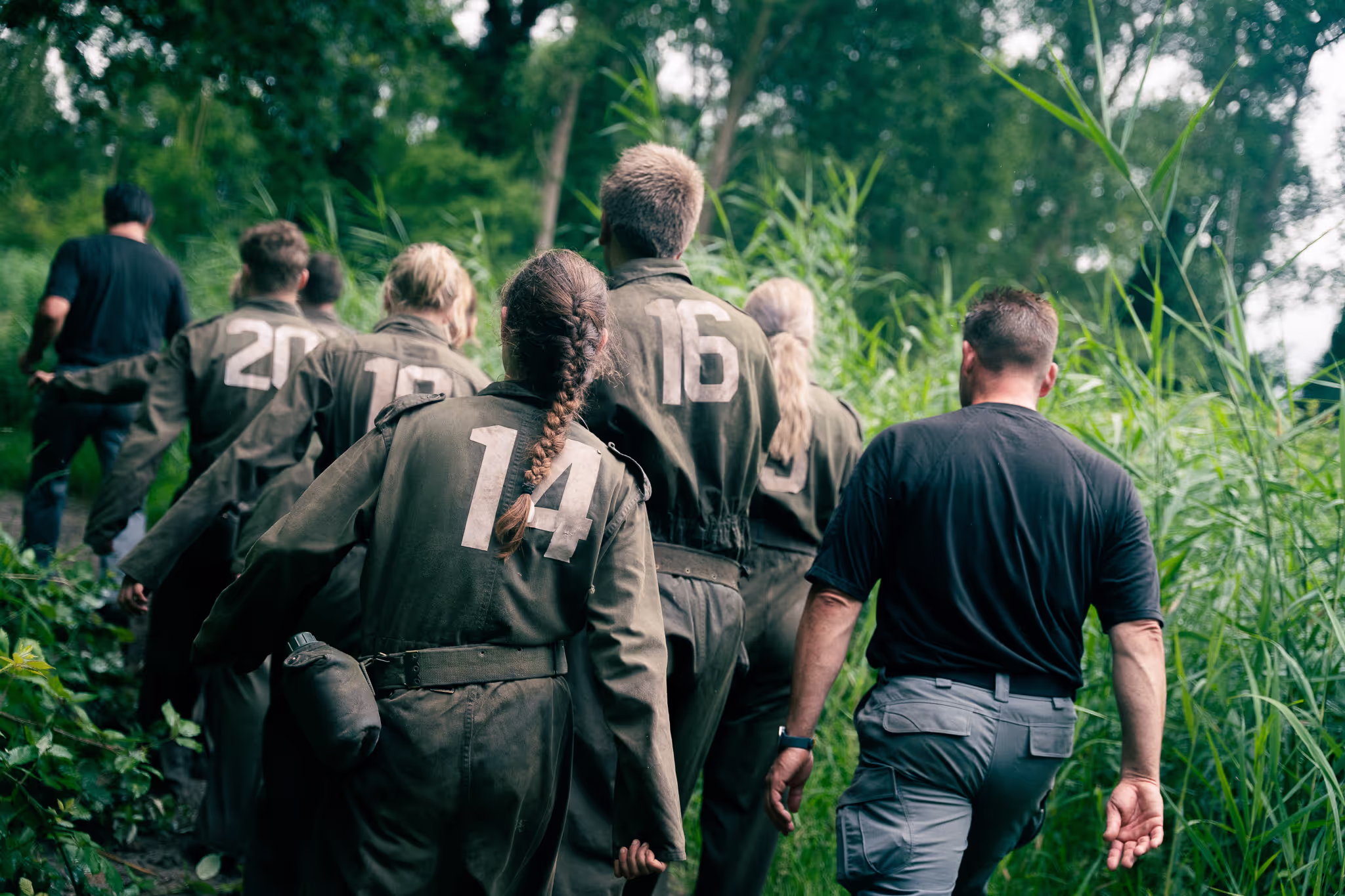 Een groep mensen in militaire uniformen met nummers op de rug, lopend door dicht groen bos.