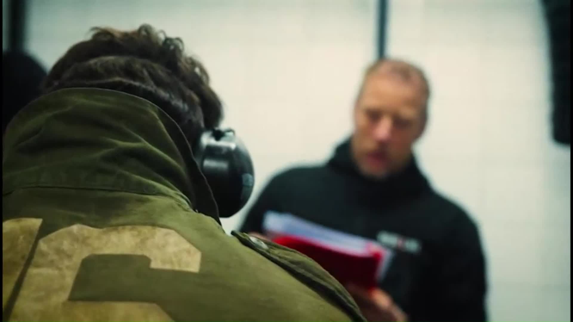 Man met donkere jas staat buiten in het donker, kijkt naar rechts met handpalmen samen.