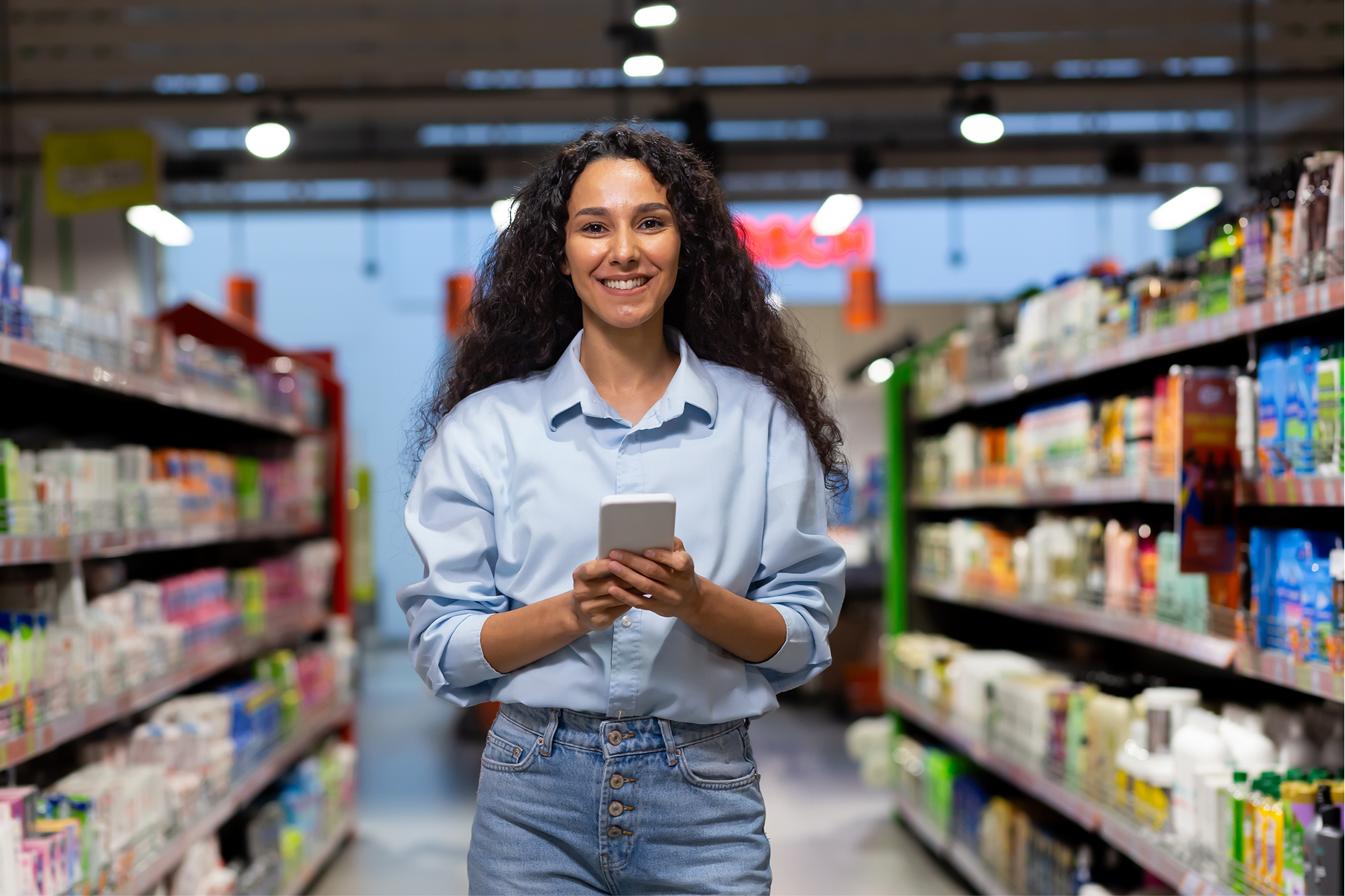 Smiling woman with curly hair using smartphone in a brightly lit store aisle with shelves of products.