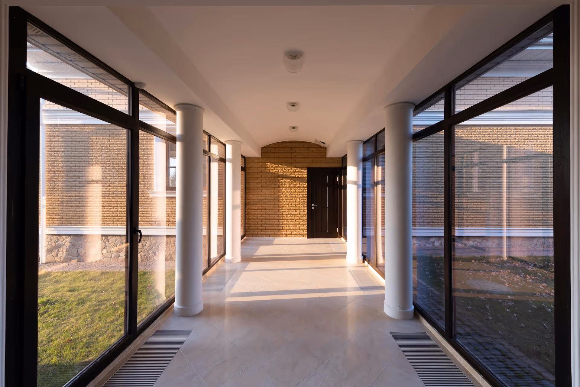 Sunlit corridor with white columns and large glass windows facing a brick wall and green lawn.