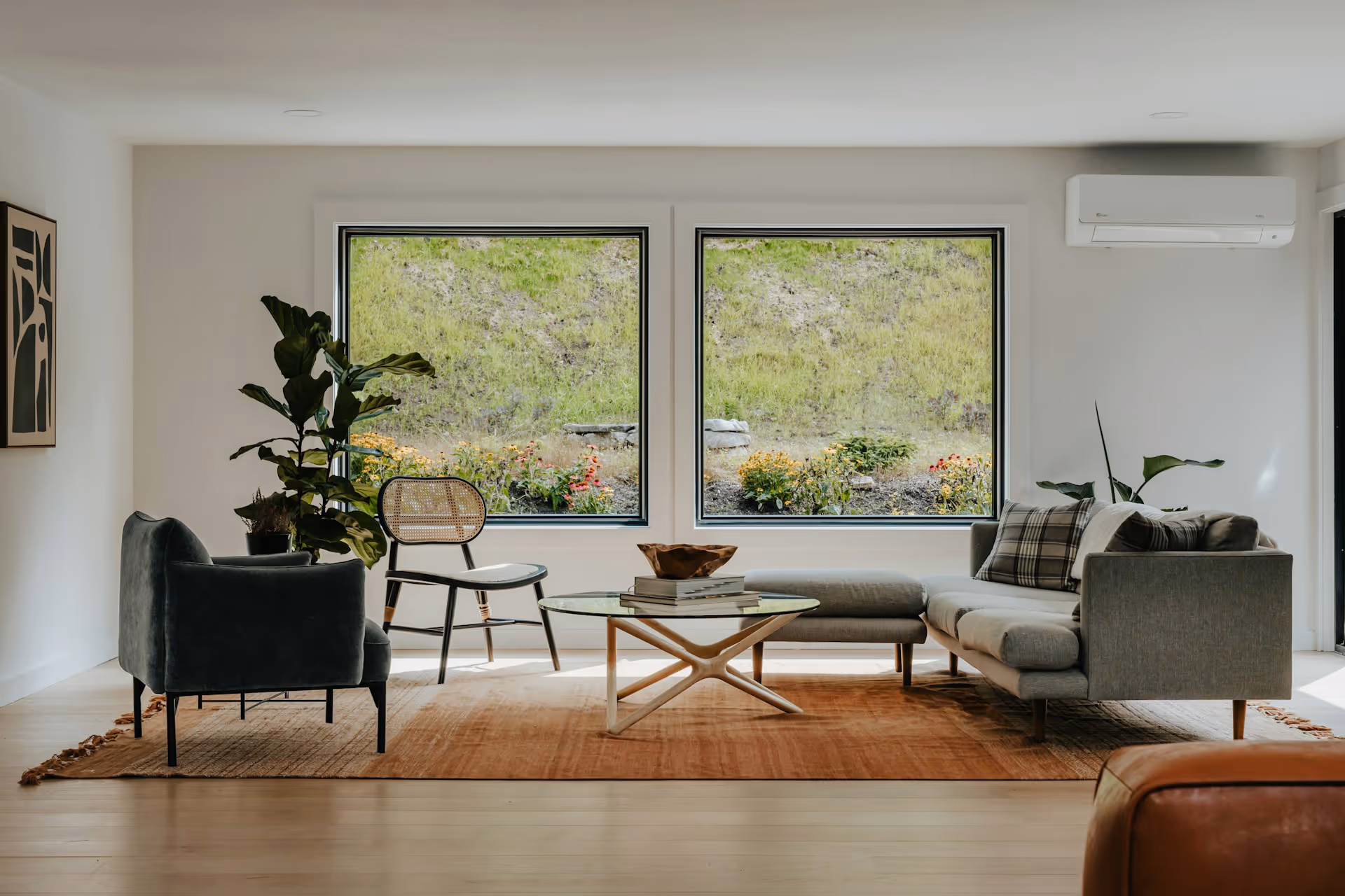 Modern living room with large windows, gray sofa, black chair, glass coffee table, and indoor plants.