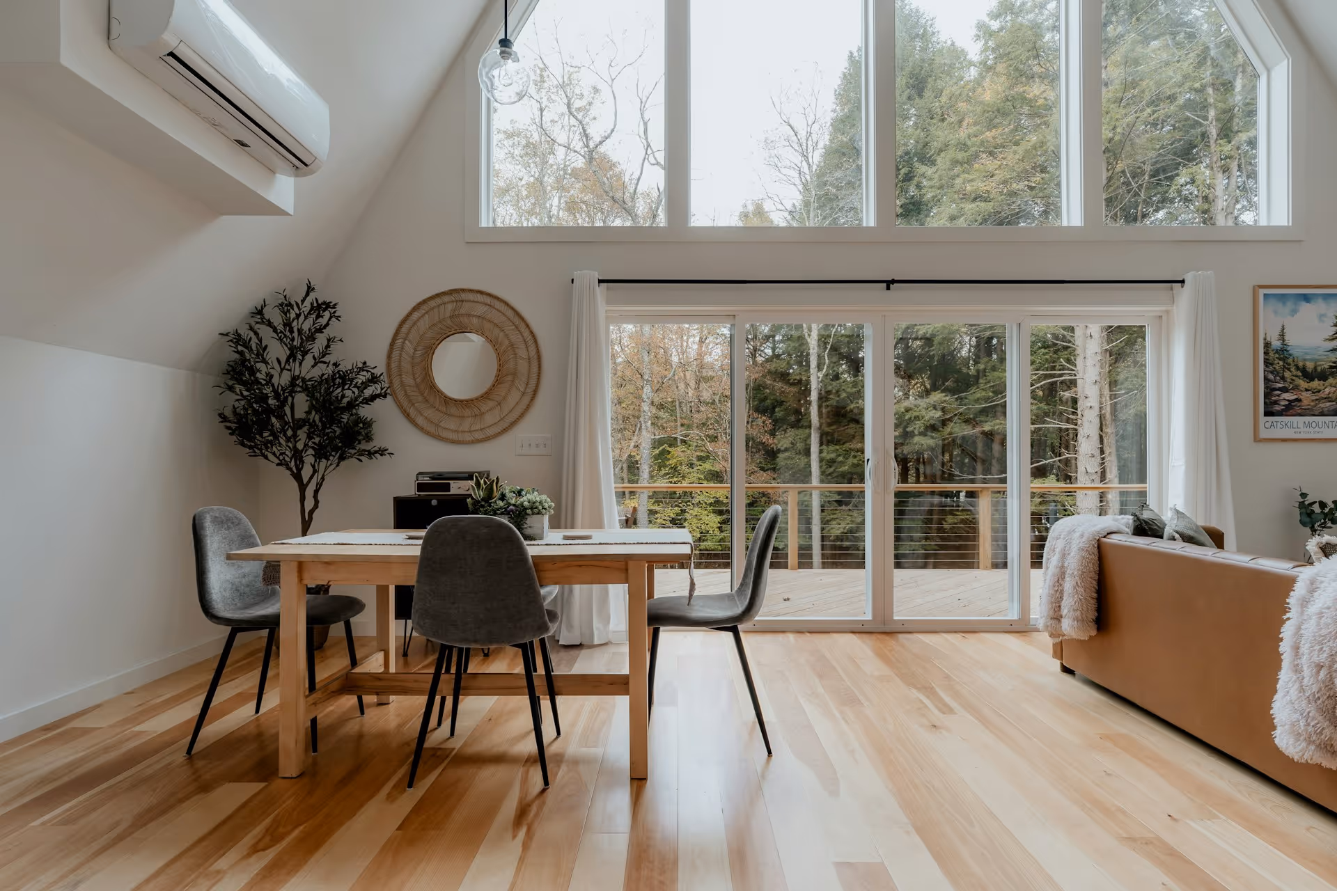 Dining area with wooden table, grey chairs, large windows, and view of trees outside on a deck.