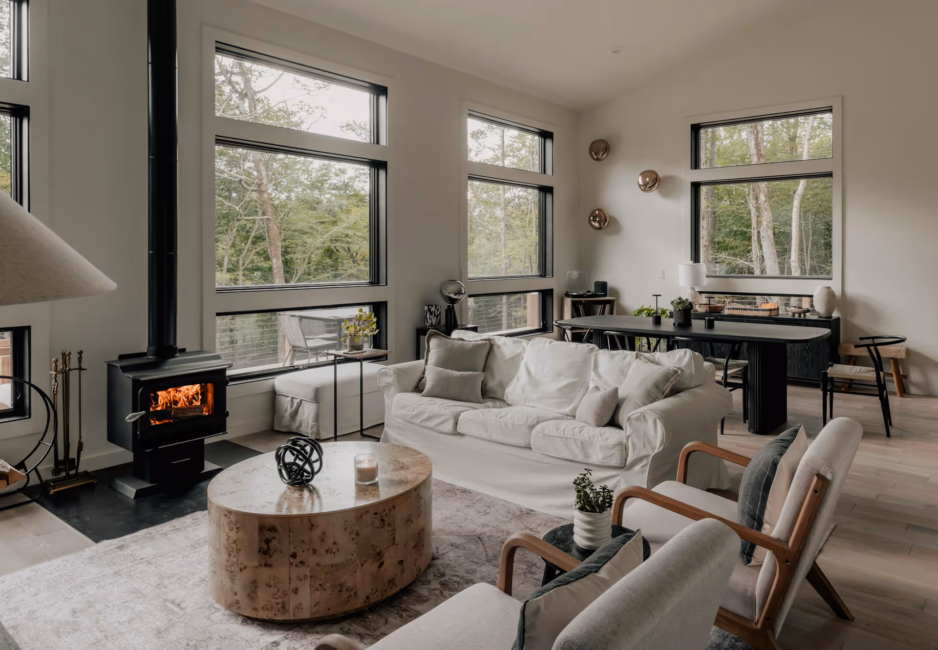 Modern living room with large windows, fireplace, white sofa, oval coffee table, and wooden chairs.