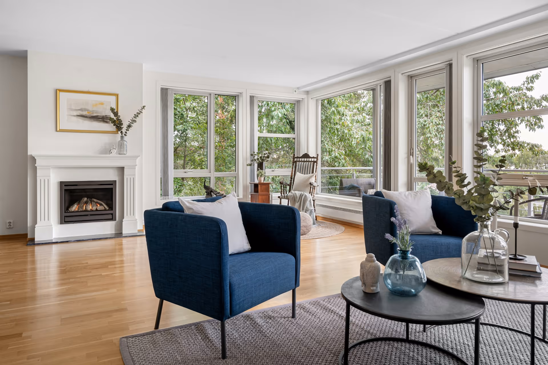 Modern living room with blue armchairs, fireplace, large windows, and wooden rocking chair near greenery view.