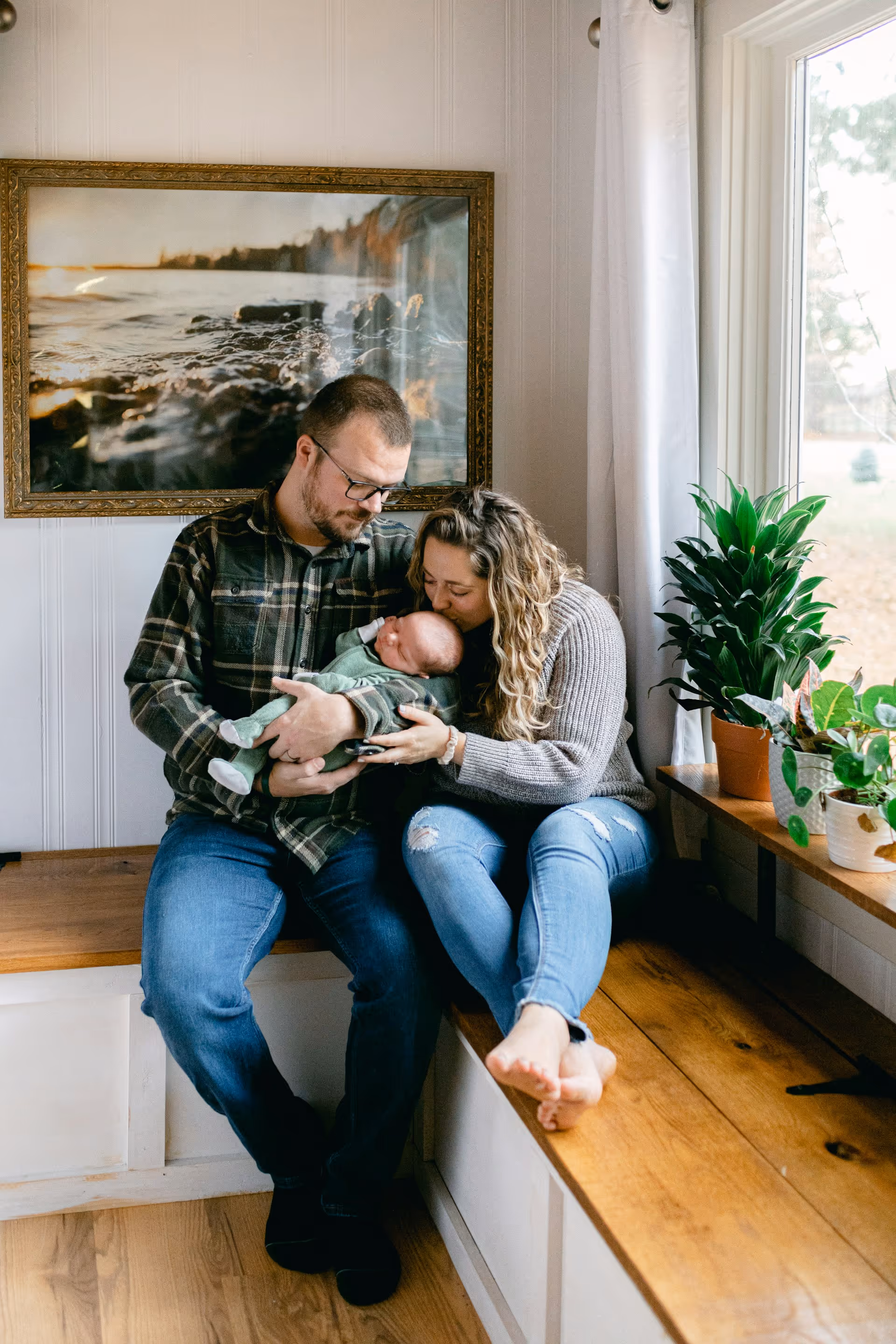 Parents sitting on a wooden bench by a window, holding and kissing their newborn baby.