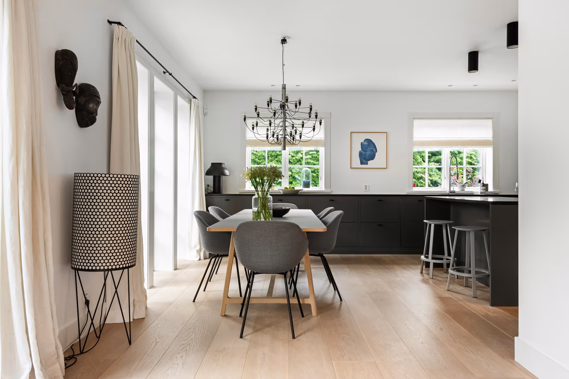 Modern dining room with a wooden table, gray chairs, a black chandelier, and windows overlooking greenery.